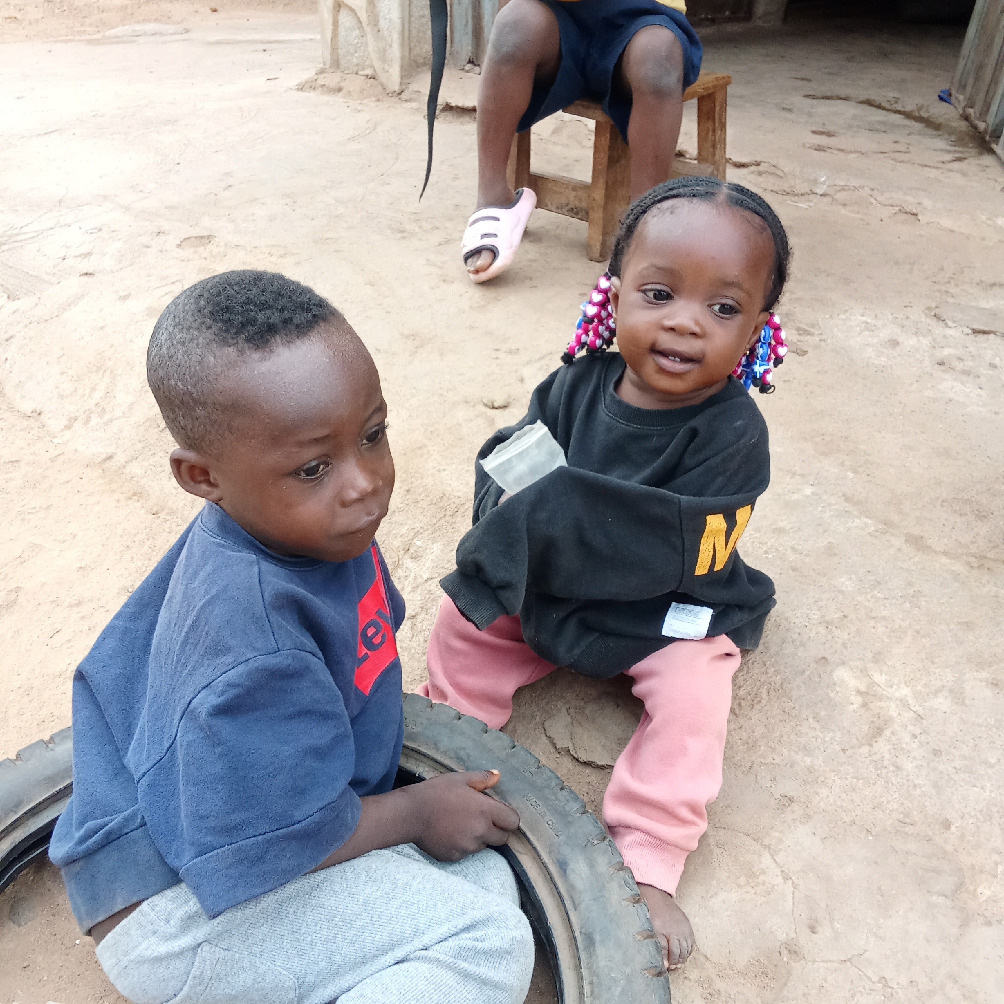 Two young children are sitting on a dirt ground in Akwanga, Nigeria. One child, wearing a blue shirt with a red design and light gray pants, is seated inside a tire, holding onto it with one hand. The other child, wearing a black sweatshirt with a yellow "N" and pink pants, sits barefoot next to the tire. The child in black is holding a small plastic bag. In the background, a person wearing blue shorts and pink sandals sits on a stool. A building is visible in the background. The ground appears dry and cracked.