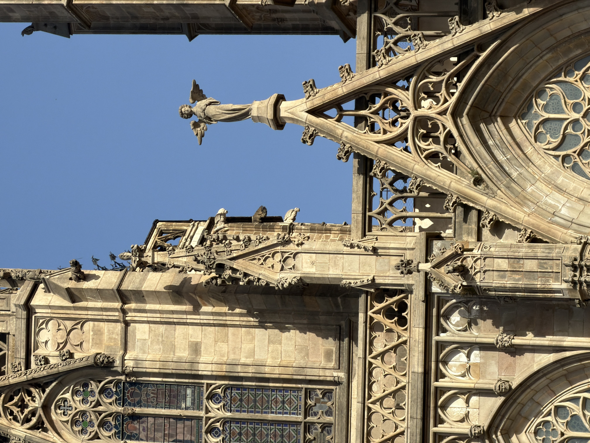 The image shows an architectural detail of a gothic cathedral with an angel statue on top of a spire. The cathedral is made of stone and features intricate carvings and stained glass windows. The sky is clear and blue. Based on the architectural style and the presence of the angel statue, this is likely a part of the Barcelona Cathedral, officially known as the Cathedral of the Holy Cross and Saint Eulalia, located in the Gothic Quarter of Barcelona, Spain. The statue is positioned high up, overlooking the city. There are no people visible in the image. The focus is on the detailed stonework and the angelic sculpture.