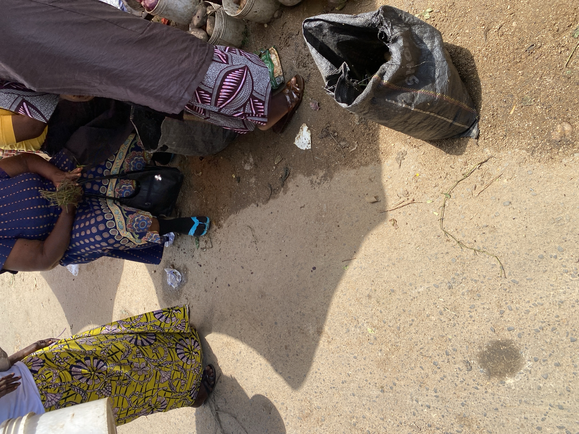 This image depicts an outdoor scene at a market in Jos, Nigeria. The ground is made of concrete and dirt. Several people are present, their lower bodies and legs visible. One person on the left wears a bright yellow skirt with a floral pattern and dark sandals. To their right, another individual is seated, wearing a blue patterned dress and blue flip-flops. A third person, partially visible on the right, wears a brown skirt with a geometric pattern and brown sandals. In the foreground, a large black plastic bag lies open. In the background, several metal buckets and bags are visible, some containing what appear to be root vegetables. The lighting suggests it is daytime, with strong shadows cast on the ground.