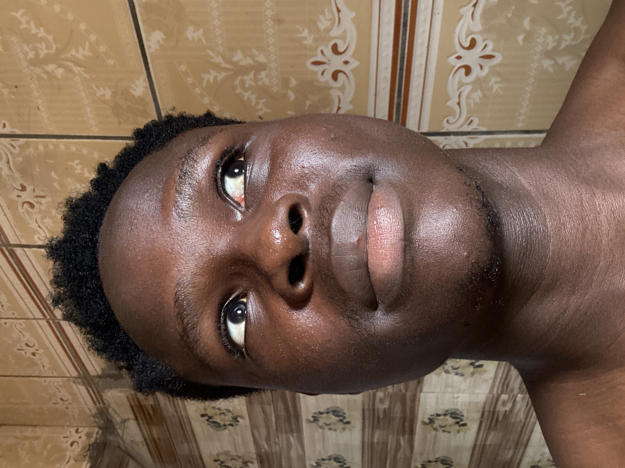 A close-up portrait features a person with dark skin and short, black, curly hair, captured at an angle with their head tilted. The individual's face, including their eyes, nose, and full lips, is prominent, with the gaze directed towards the camera. A slight reddish tint is visible in the inner corner of one eye. The skin exhibits a noticeable sheen, reflecting light, particularly on the forehead, nose, and lips.
The background consists of patterned ceramic tiles in shades of tan and beige, adorned with elaborate brown and off-white floral or geometric designs. These patterns suggest an indoor setting, likely a wall. The tiles are arranged in vertical and horizontal lines, forming a decorative backdrop.
The activity depicted is a simple portrait or selfie, focusing on the individual's appearance. The lighting appears to be artificial, creating highlights on the skin. No specific event is taking place beyond the act of being photographed. The image does not contain any visible text. This scene is situated in Ihiagwa, Nigeria.