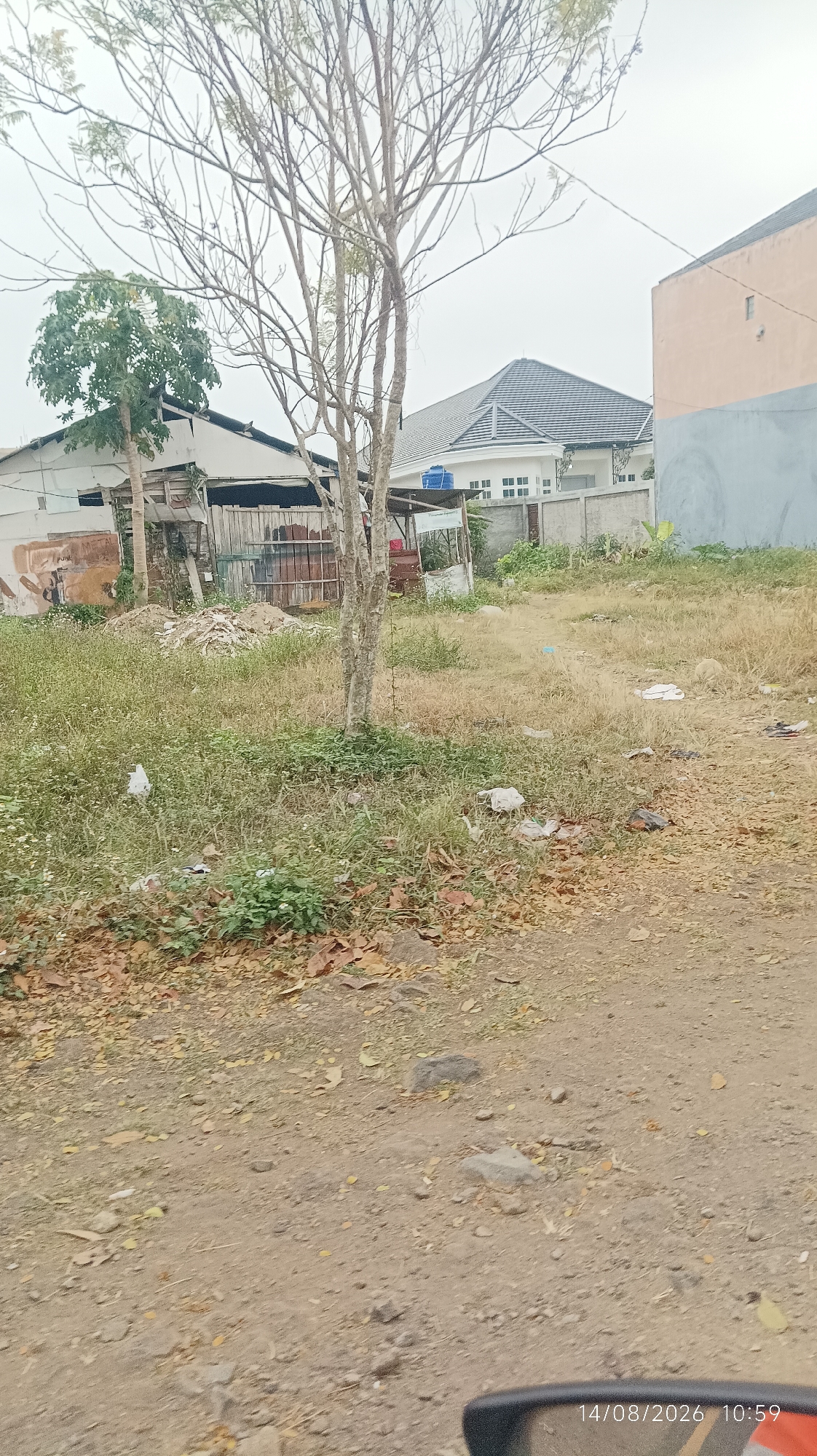 The image depicts a cluttered vacant lot with overgrown grass and scattered debris, including plastic bags and leaves. In the background, there are several buildings. On the left, there is a dilapidated structure with a corrugated metal roof and wooden supports. Next to it, a two-story house with a gray roof and white walls is visible. To the right of this house, a modern building with a gray tiled roof and white facade stands behind a concrete fence. The ground is a mixture of dirt and dry grass, with several rocks scattered about. The scene appears to be located in an urban or semi-urban area, given the presence of multiple buildings. The timestamp at the bottom right corner indicates the image was taken on August 14, 2026, at 10:59. Based on the characteristics of the buildings and vegetation, this scene is consistent with a location in the city of Tasikmalaya, Indonesia.