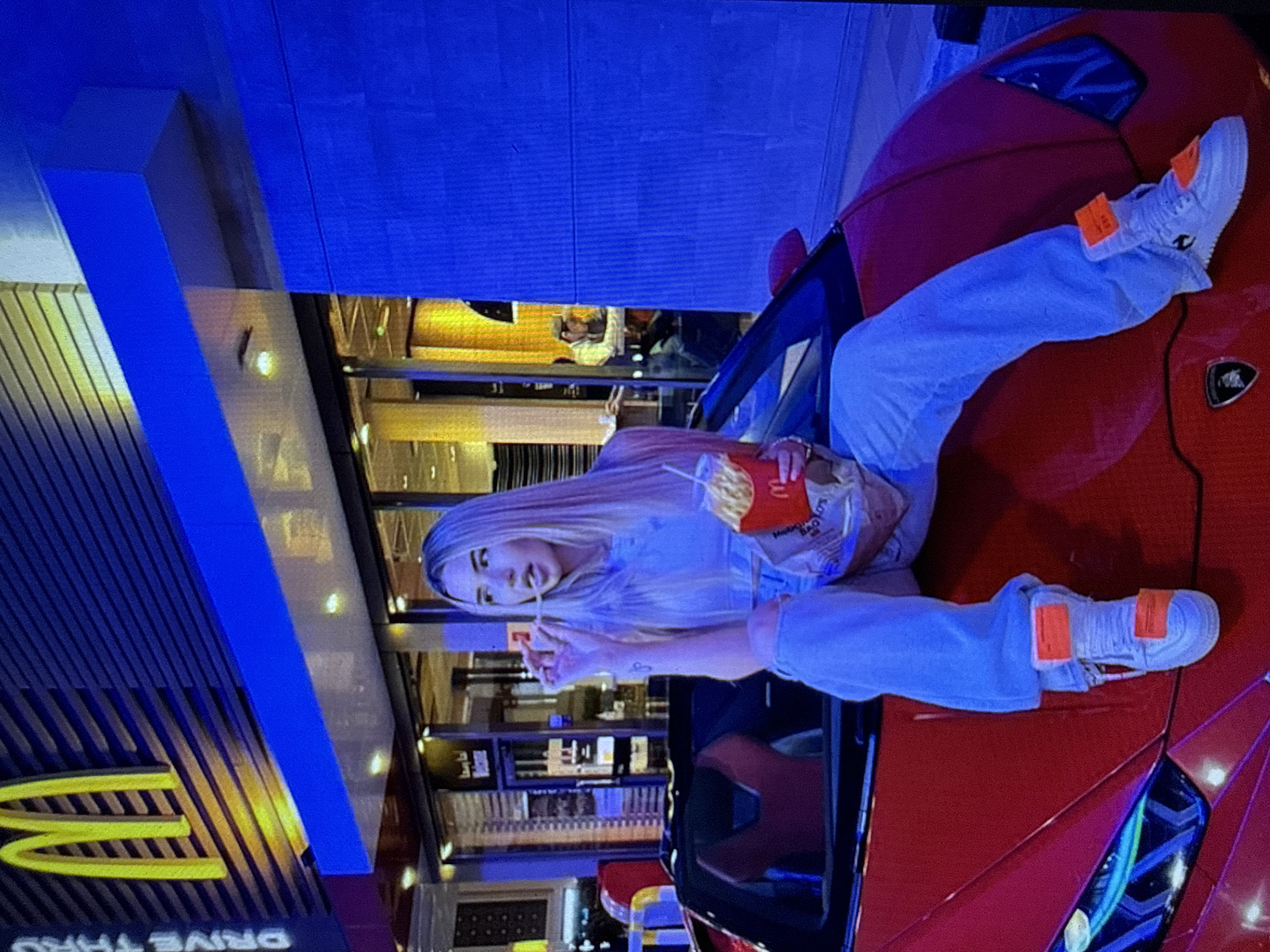 The image, reportedly taken in Kardzhali, Bulgaria, depicts a nighttime scene in an urban environment. A young woman with long, straight blonde hair and fair skin is prominently featured, seated atop the red hood of a Lamborghini convertible. She is dressed in a light-colored crop top, light blue ripped jeans, and white sneakers accented with bright orange tags or overlays. Her left hand holds a McDonald's branded French fry container and a drink cup, while her right hand holds a single French fry near her open mouth, indicating the act of eating. A small, dark-colored tattoo is visible on her left forearm.
In the background, a McDonald's restaurant is clearly identifiable by its illuminated yellow "M" logo on a dark blue facade. The words "DRIVE THRU" are partially visible on the left side of the building. The exterior of the restaurant and the surrounding area are lit by a prominent blue artificial light source, contrasting with the warm yellow interior lighting visible through the large windows. One individual is partially discernible through the restaurant's window on the right side, appearing to be seated at a table. The red Lamborghini features distinct blue and green LED headlights and a visible black Lamborghini emblem on its hood. The vehicle's top is down, revealing dark interior seating with red accents.