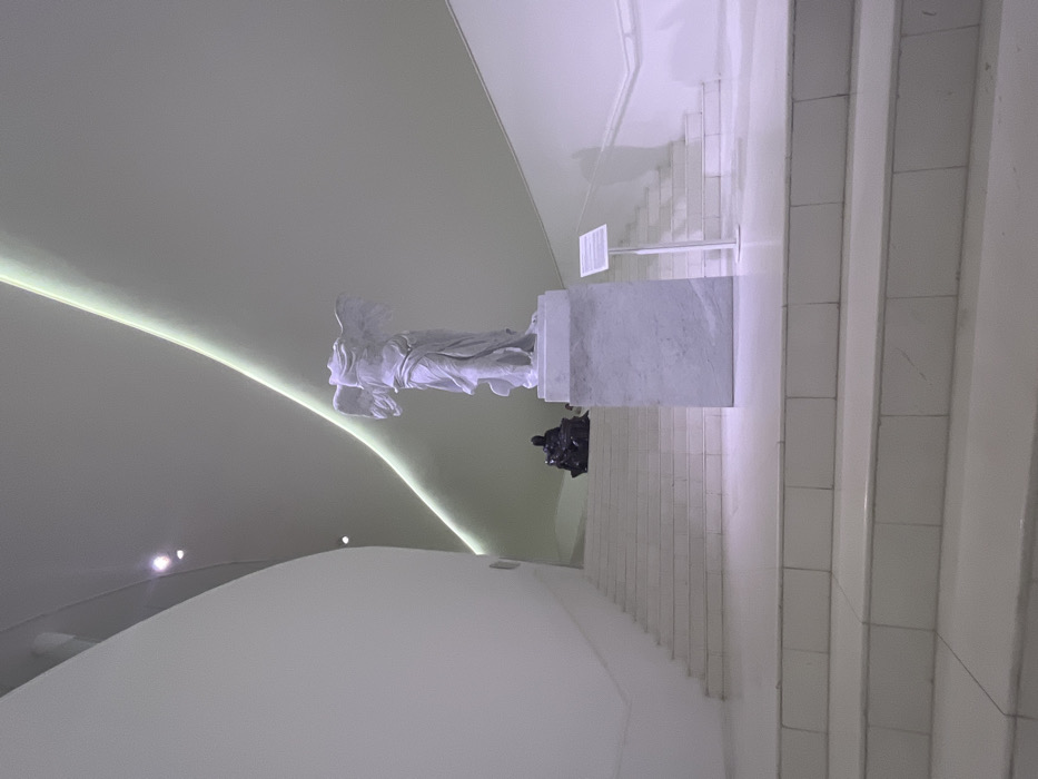The Winged Victory of Samothrace statue sits atop a marble base in a bright, modern museum. The walls are curved and painted white, with a thin, glowing light strip above the statue. A black, smaller statue of a seated person is also present, next to the larger sculpture. White stone stairs line the right side of the image, going up and away. A white sign stands next to the statue, likely containing information about the exhibit.