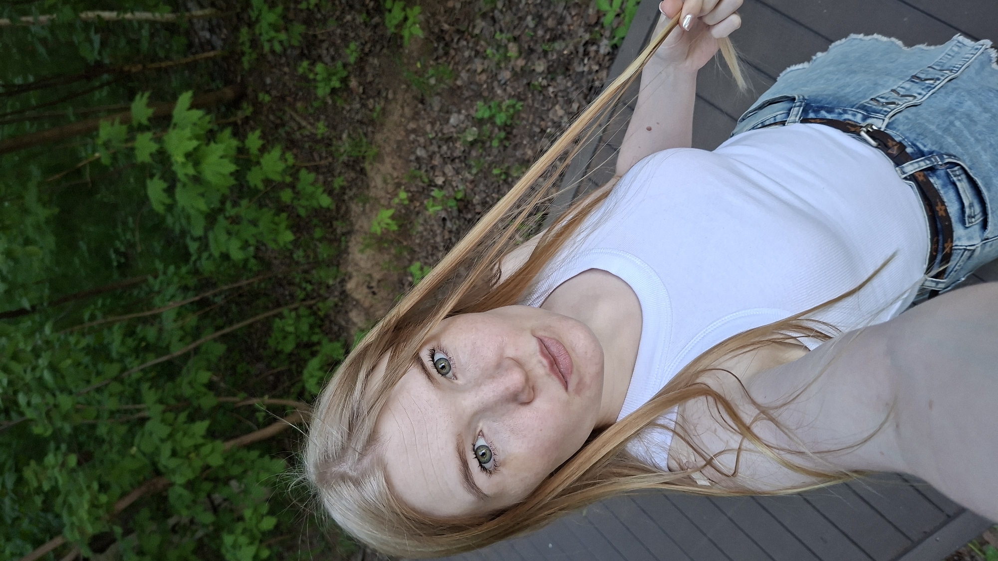 A woman with fair skin and long blonde hair, wearing a white tank top and denim shorts with a brown belt, is looking directly at the camera. The background includes green foliage suggesting a natural environment like a forest or park. The ground appears to be wooden planks, possibly a deck or boardwalk. Given the prompt mentioning the city of Chaikovsky, Russia, it is reasonable to infer that the photo was taken in or near a natural area within or surrounding Chaikovsky.