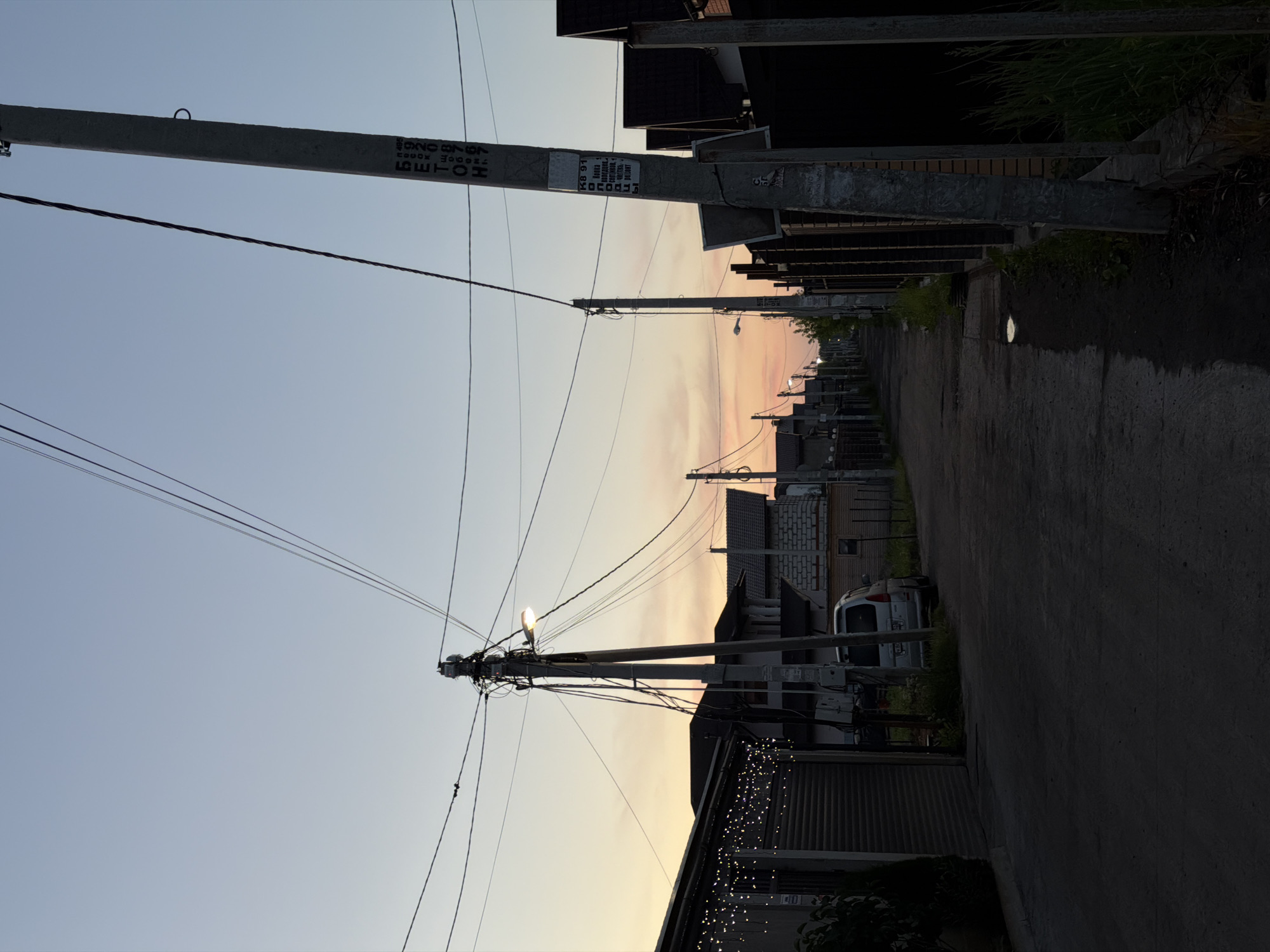 A rural street is depicted at dusk, with a clear sky transitioning from light blue to a soft pink and orange hue near the horizon. The street is unpaved and shows signs of wear, with puddles reflecting the dim light. Power lines crisscross the sky, supported by concrete poles. Houses with peaked roofs line both sides of the street. A silver minivan is parked on the right side of the street. The left side of the street features a building with a corrugated metal garage door adorned with a string of small, bright lights. A concrete pole on the right side of the street has various markings and stickers, including what appears to be Russian text. The overall atmosphere is quiet and peaceful, suggesting a suburban or rural residential area.