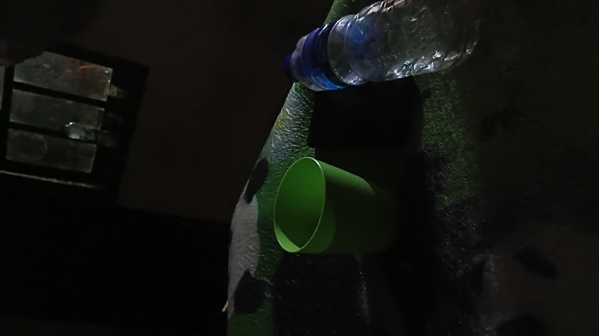 The image shows a green plastic cup and a partially filled plastic water bottle on a dark, textured surface that appears to be a mat or fabric with a camouflage pattern. In the background, a dimly lit window with multiple panes is visible. The overall scene is dark, suggesting it might be nighttime or an indoor environment with low lighting. There is no indication of the specific location in Prabumulih, Indonesia, or any activity involving people.