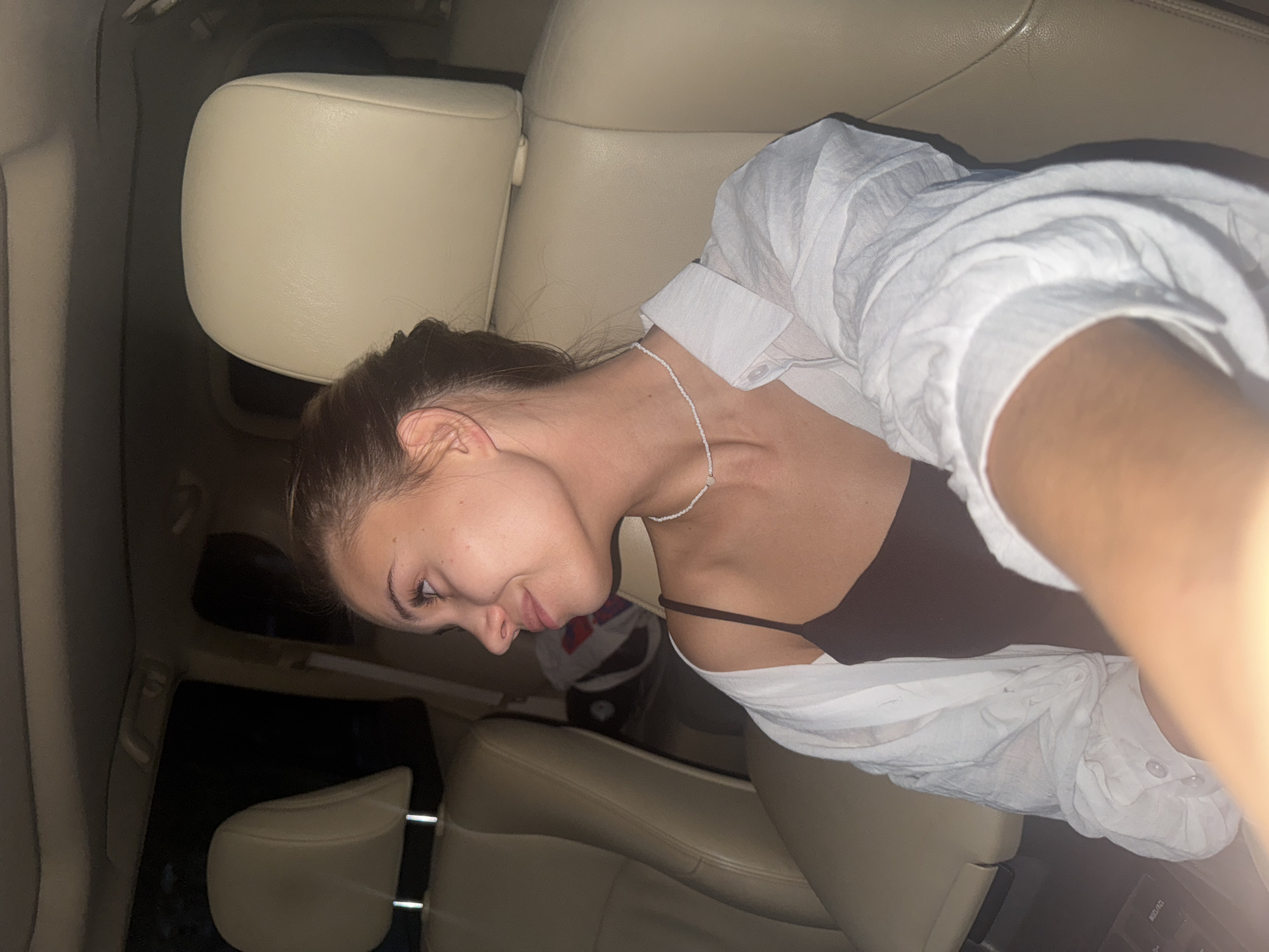 A young woman is sitting in the passenger seat of a car, wearing a white button-down shirt over a brown tank top and a white beaded necklace. The interior of the car features beige leather seats. The location is likely Chaikovsky, Russia, but specific landmarks or identifiers within the image do not confirm this. There are no visible actions or interactions taking place. The surrounding environment suggests it is nighttime or a dimly lit area.