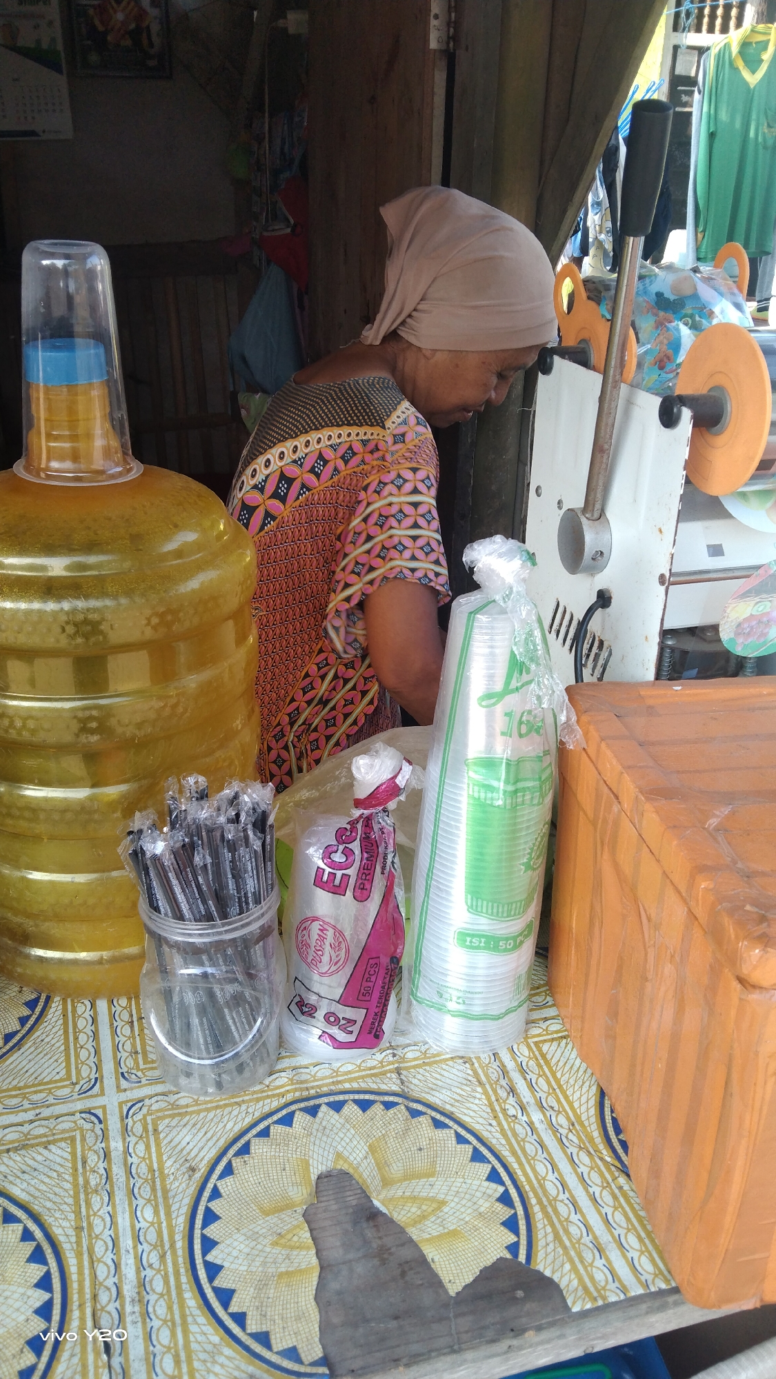 The image captures a scene at a roadside stall in Karawang, Indonesia, likely a beverage or convenience stand. In the foreground, a large yellow water dispenser dominates the left side of the frame, topped with a clear plastic cup containing a blue lid. To its right, a woman wearing a light brown headscarf and a vibrantly patterned pink and orange shirt is partially visible, facing away from the camera and looking down. She appears to be tending to a sealing machine, a portion of which is visible to her right.
In front of the woman and the water dispenser, a collection of disposable plastic cups is arranged. Two stacks of clear plastic cups, one larger and one smaller, are clearly visible. The larger stack has green printing on it. To the left of the cups is a clear plastic container filled with individually wrapped black straws. A partially visible orange cooler box is situated to the right of the plastic cups.
The stall appears to be an open-air setup, suggested by the visible wooden structures and hanging clothing in the background. The surface where the items are placed is covered with a patterned tablecloth featuring circular designs. The overall impression is one of a bustling, everyday scene at a local business in Indonesia.