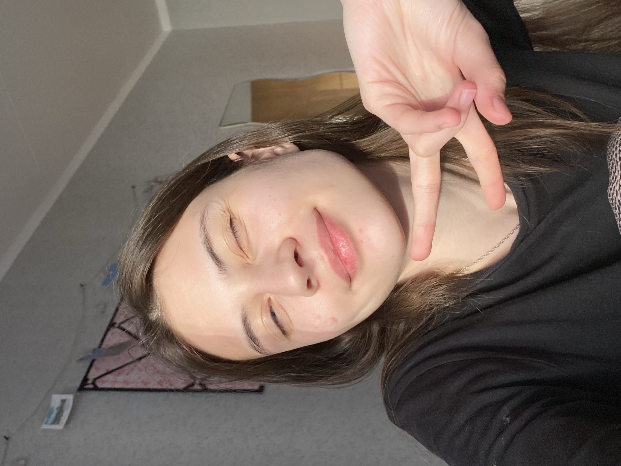 A young woman with brown hair is positioned at an angle, facing the camera. Her right hand is raised in a peace sign, partially obscuring her face. She is wearing a black top and a delicate silver chain necklace. The background includes a gray wall, a rectangular mirror, a picture in a frame, and a decorative piece. The location appears to be indoors, possibly a bedroom or living space. There is no information available that indicates this photo was taken in Votkinsk, Russia.
