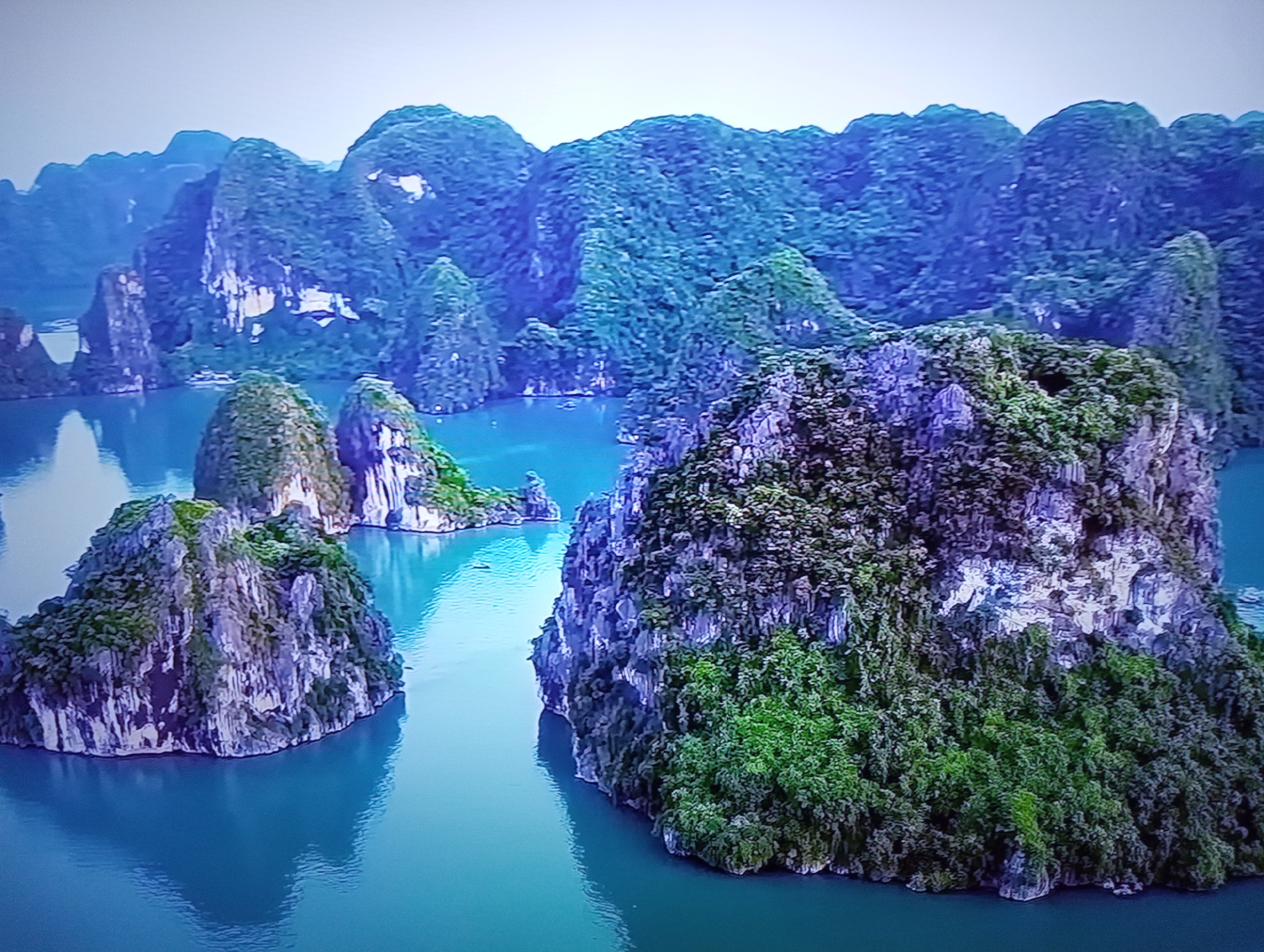 This media file depicts a scenic aerial view of a karst landscape with numerous limestone karsts rising from a body of tranquil, turquoise water. The karsts are covered in lush green vegetation, creating a dramatic contrast with their rocky, weathered surfaces. In the foreground, a large karst with dense foliage dominates the view, while other karsts of varying sizes are scattered throughout the water, some appearing closer and others further in the distance. The background features a hazy mountain range, also covered in vegetation, stretching towards a pale, overcast sky. The overall scene suggests a natural, possibly tropical, environment. There are no visible people, objects, or text. The time of day appears to be daytime, with the overcast sky indicating diffused lighting and possibly a calm or slightly hazy weather condition. Although the prompt provides a location context of Dnipro, Ukraine, the visual elements of the image are not consistent with this location and strongly suggest a tropical island setting, such as Halong Bay in Vietnam.