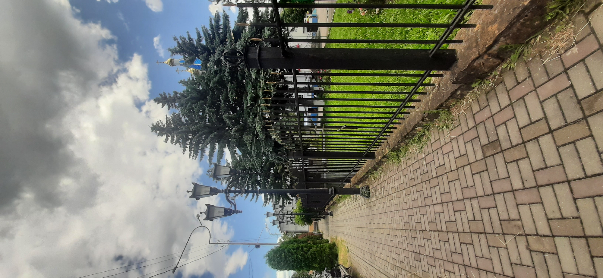 The image depicts a paved pedestrian walkway with brick-like pavers, extending into the distance on the left. A black metal fence with vertical bars and decorative finials runs along the right side of the walkway. Beyond the fence, a strip of bright green grass is visible, with some dry grass and weeds growing at the base of the fence. In the background, several tall, dark green evergreen trees are present, with the golden domes and blue roofs of a church visible behind them. The sky above is a mix of bright blue with scattered white and dark gray clouds. Several ornate black lampposts with multiple lamps are positioned along the walkway. The scene appears to be in a park or near a public building.