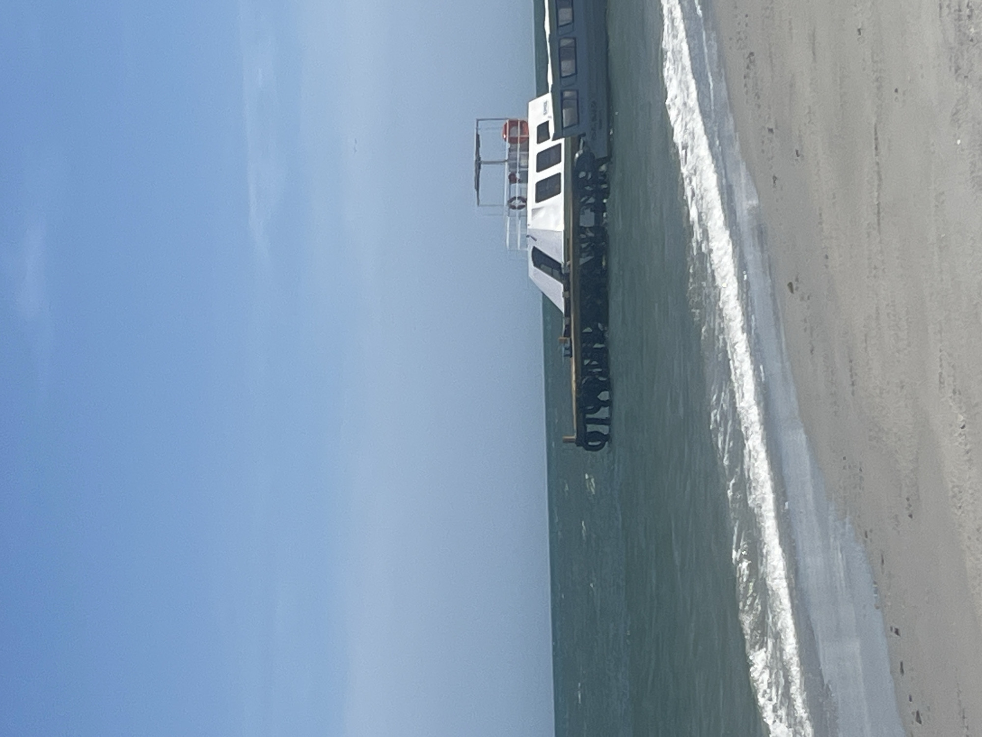 The image depicts a motor boat docked next to the beach in El Yaque, Venezuela. The boat is white with black accents and sits next to the beach, with a clear blue sky overhead. The sand is light in color and there are small waves washing ashore. The boat has several windows and a ladder on top.
