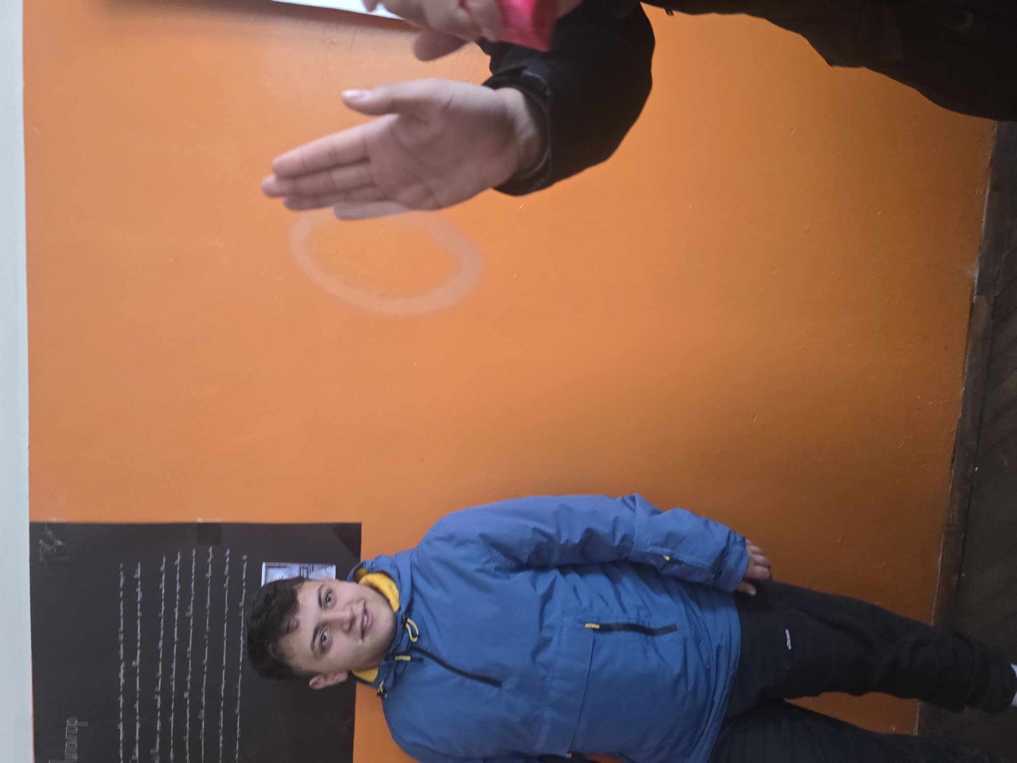 The scene depicts an indoor environment with two individuals against an orange wall.
**Environment:** The background is a vibrant orange wall. To the upper left, a black poster board, approximately rectangular, displays white text in a language resembling Bulgarian and includes smaller photographic inserts. A subtle, light-colored circular marking is visible on the orange wall to the right of the center. The floor appears to be dark wooden planks with a dark baseboard at the wall's bottom. A white trim runs along the top edge of the orange wall.
**People:**
1. **Subject 1 (Left):** A young male, appearing to be in his late teens or early twenties, stands on the left side of the frame. He has short, dark hair and is looking slightly to his right with a faint smile. He is dressed in a blue hooded anorak jacket featuring a visible yellow lining around the neck and yellow zipper accents on pockets. He wears dark athletic trousers with a white 'ellesse' logo on the left thigh.
2. **Subject 2 (Right):** Only the right arm and hand of a second individual are visible on the right side of the frame. The hand is raised, palm open, facing towards the left. This individual is wearing a black long-sleeved garment. A small, bright pink object is partially visible, clutched in their left hand, at the extreme right edge of the image.
**Actions/Interactions:** The male on the left is standing in a relaxed posture, observing the scene to his right. The partial figure on the right is engaged in a gesture with a raised open palm, potentially indicating a stop, a wave, or part of a dynamic interaction.
**Location:** The specific location within the city of Samokov, Bulgaria, such as a particular building or address, cannot be determined from the visual information provided. The setting, with its poster and wall, suggests a public or institutional indoor space, such as a school, community center, or event venue.