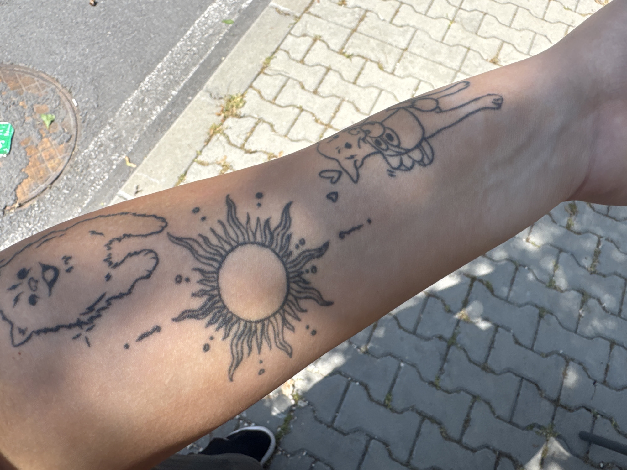 The image shows an arm with three black ink tattoos. One tattoo is of a cat with a crescent moon. Another tattoo is of a sun with rays emanating from its center. The third tattoo depicts another cat lying down with its back to the viewer. The tattoos are arranged linearly along the forearm. The environment includes a brick or stone walkway and a glimpse of what appears to be a street with a manhole cover. A person wearing what appears to be a shoe is also visible in the image.