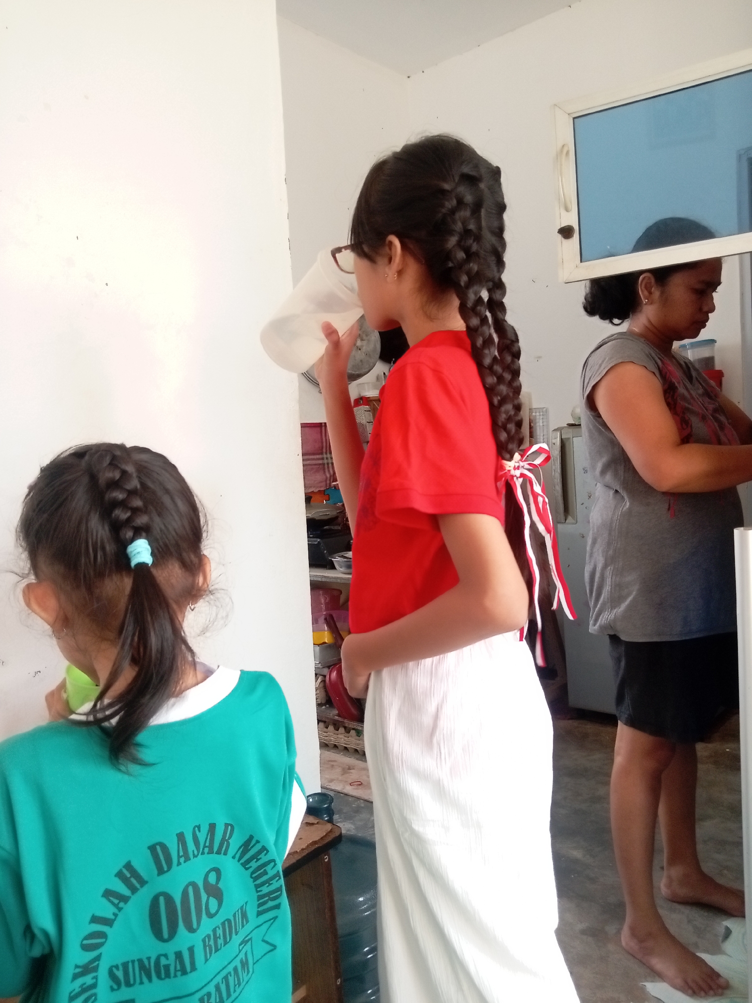 In an interior scene that appears to be a kitchen or utility area in Batam, Indonesia, three individuals are present. In the foreground, a young girl with dark hair styled in two braids is drinking from a translucent plastic cup, holding it with her right hand. She is wearing a red t-shirt and a white, pleated skirt. To her left and slightly further back, another young girl faces away from the camera, also with dark hair in a braid, wearing a teal t-shirt with text printed on the back. Behind these two, a woman with dark hair pulled back stands to the right, wearing a grey t-shirt and dark shorts. She is looking down and appears to be engaged in an activity involving food or dishes. The environment includes a white wall, a window with a blue tint, a metal cabinet or refrigerator, and shelving with various containers. The floor is a light-colored, possibly concrete or tiled surface. The overall impression is a candid moment captured within a domestic or communal space.