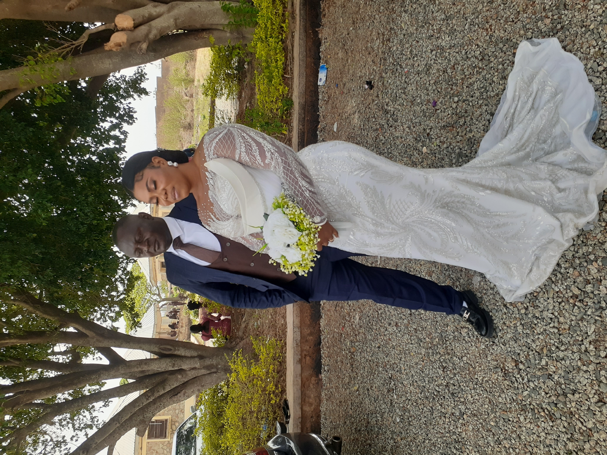 A photograph shows a bride and groom posing for a photo on a gravel path in Unknown, Unknown. The bride is wearing a white, long-sleeved wedding dress with lace detailing, holding a white bouquet and looking at the groom. The groom is wearing a navy suit with a brown vest and tie. The setting appears to be a park or outdoor event space with trees and a building visible in the background. Other individuals and a car are partially visible in the background. A piece of trash is on the ground.