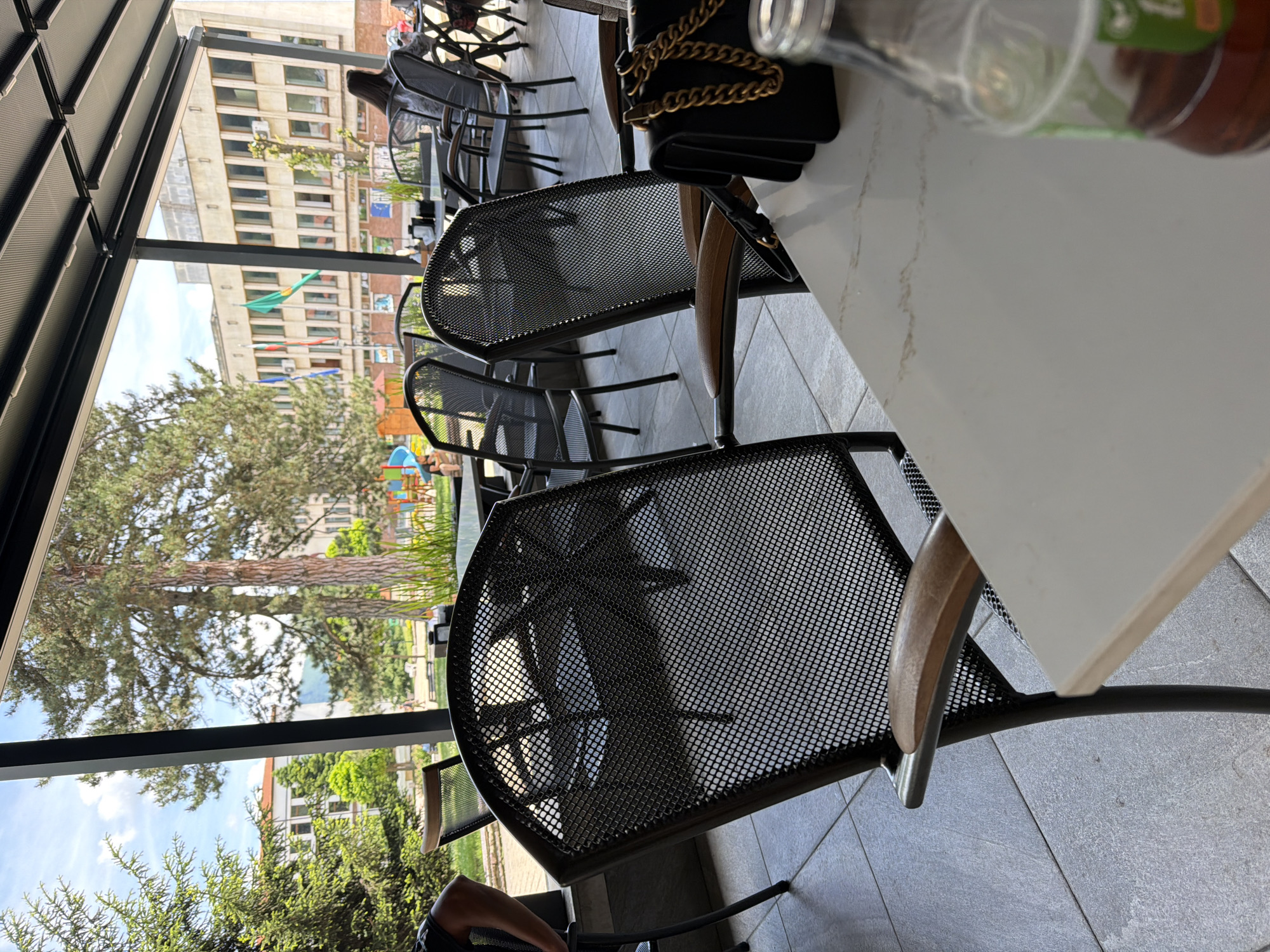 The image depicts an outdoor seating area of a cafe, likely in Etropole, Bulgaria, with a large building featuring multiple windows and flags visible in the background. In the foreground, several dark metal chairs with wooden armrests are arranged around tables. A black handbag with a gold chain rests on one of the tables, next to a clear glass bottle containing a brown beverage. The ground is paved with large square tiles. Trees and a playground are visible in the middle ground, suggesting a public space. The overall atmosphere is that of a relaxed daytime setting.