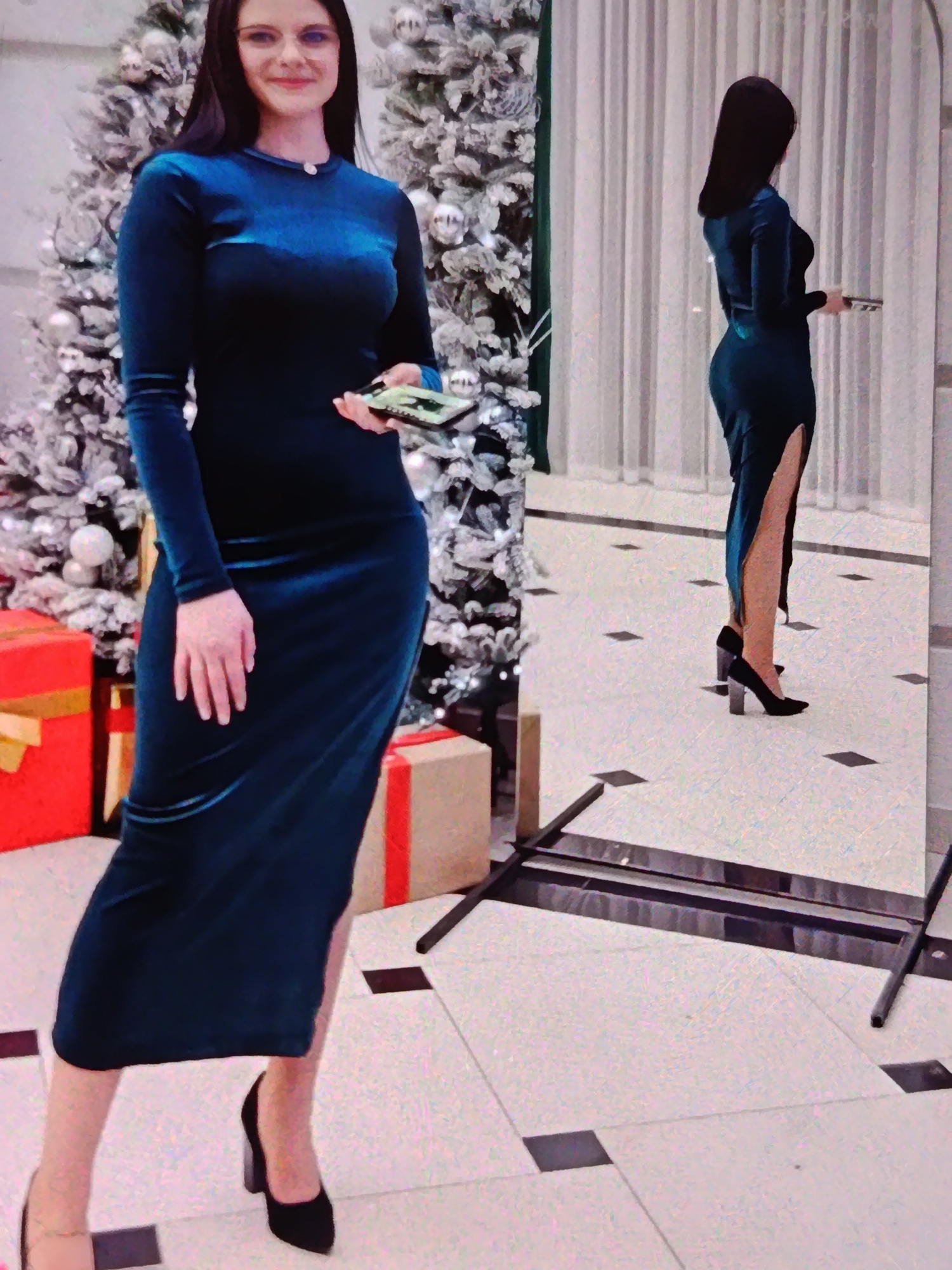 A woman stands in front of a decorated Christmas tree and a large mirror, holding a cell phone. She is wearing a long-sleeved, dark teal velvet dress with a high slit up the side and black high-heeled shoes. The Christmas tree is adorned with white and silver ornaments, and red gift-wrapped boxes are visible in the foreground. The woman is smiling, with long dark hair and glasses. The scene appears to be indoors, possibly a home or event space, with tiled flooring and curtains in the background. A reflection of the woman in the mirror shows her from the side, highlighting the dress's silhouette and the slit. The lighting suggests it could be daytime or evening. There is no visible text in the image.