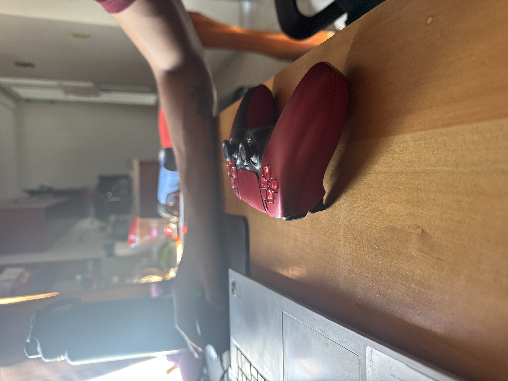 The image shows a red PlayStation 5 DualSense controller sitting on a wooden desk. A person's arm is visible near the controller. A laptop is partially visible below the controller. The background is blurry, but it appears to be a room with various objects. There is no information present in this image to indicate the location is San Andrés Cholula, Mexico.