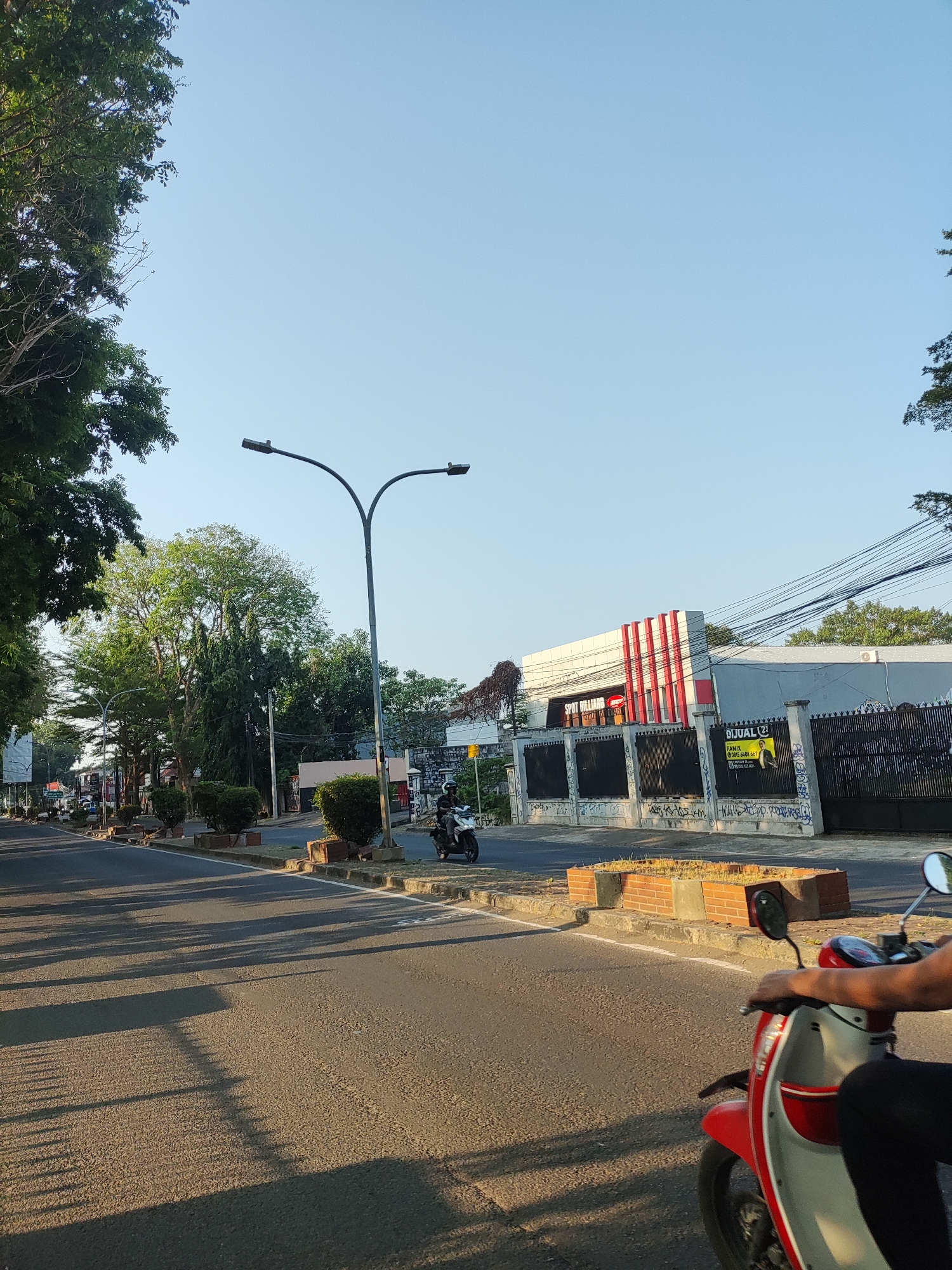 This image depicts a street scene in Cirebon, Indonesia. On the right side of the road, a red and white scooter is being ridden by an unseen person whose arm and hand are visible, gripping the handlebars. Further down the street, a person on a white motorcycle is also visible. The street is lined with trees and planters on both sides. A building with red and white striped accents is visible behind a black metal fence, with a yellow "DIJUAL" (For Sale) sign posted on the fence. Utility poles and wires are present overhead. The sky is clear and blue, indicating daytime. The road is paved and appears to be an urban or suburban area.