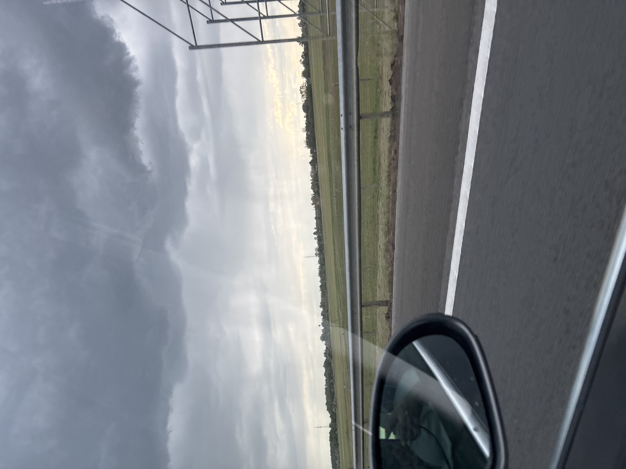 The image depicts a view from a car traveling on a highway. To the left of the road, a grassy field stretches towards the horizon under a cloudy sky. A guardrail runs parallel to the road, separating it from the field. The side mirror of the car is visible in the lower right corner. A white lane marking is present on the road. The location is near Balástya, Hungary, based on similar landscapes and infrastructure found in the area. There are no people visible. The scene is captured from a moving vehicle.