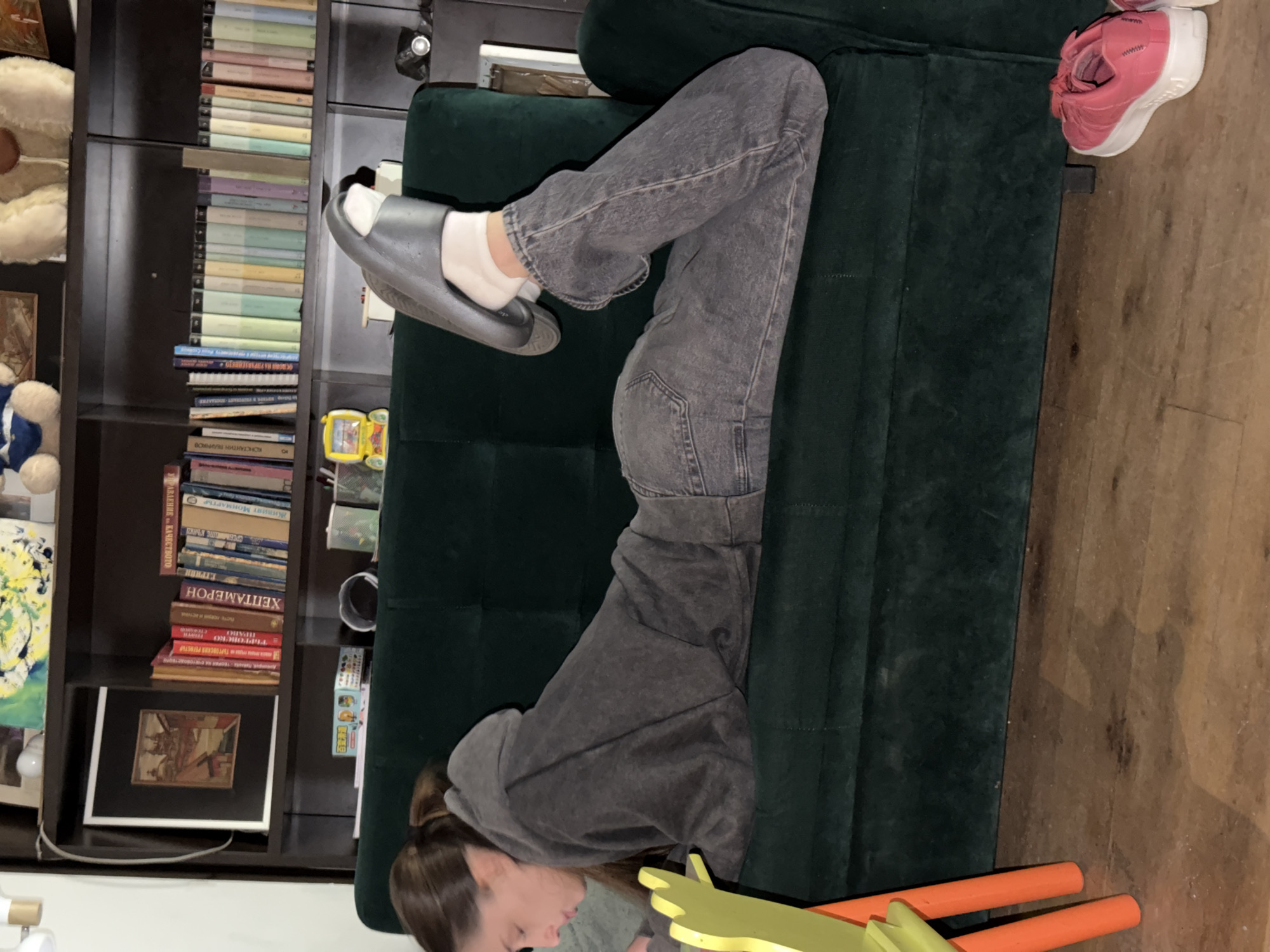 A person in a grey hoodie and jeans is lying on a green couch, with their feet propped up on the couch's back. They are wearing a grey slide on their left foot with white socks. To the left, there is a dark brown bookshelf filled with books, a stuffed bear, and other objects. To the right, there is a pink shoe on the couch. Below the couch are the legs of a yellow and orange chair. The location appears to be indoors, possibly in Sofia, Bulgaria, but the specific location cannot be confirmed from the image.