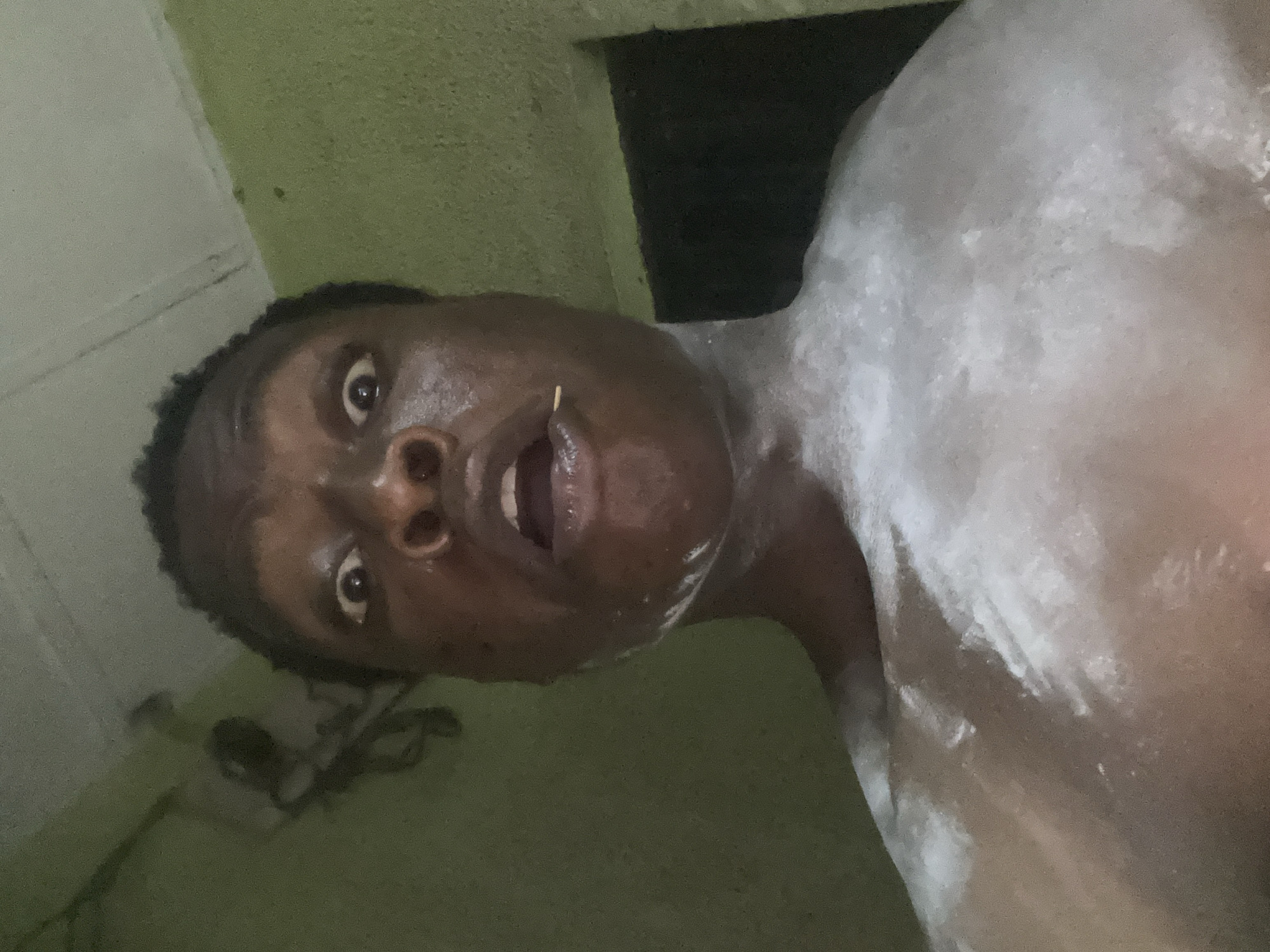 A close-up, low-angle photograph shows a shirtless person with dark skin and short, dark hair looking up at the camera. The person appears to be covered in a white, powdery substance on their chest and shoulders. The background is indistinct, showing what appears to be a green-painted wall. There is a small, thin object, possibly a toothpick, protruding from the person's mouth. The scene appears to be indoors, the location is in the city of Unknown, Unknown.