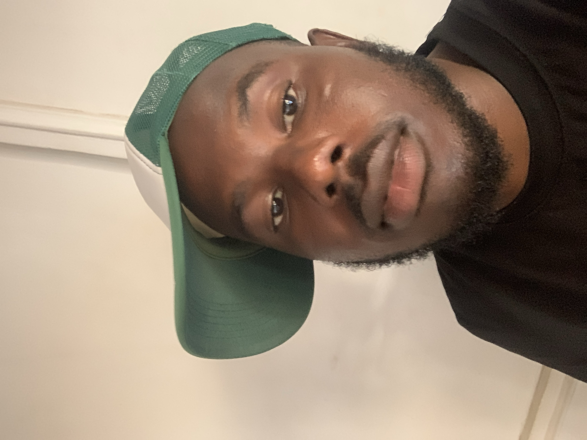 The media file presents a close-up portrait of a man with dark skin, looking directly into the camera. He is seen at an angle, with the image rotated 90 degrees counter-clockwise. The man wears a green and white baseball cap, characterized by a solid green brim and mesh back, and a white front panel. He is also dressed in a plain black t-shirt. The man has a well-trimmed beard and mustache. His expression is neutral, with a slight hint of a smile, and his eyes appear bright due to reflections. The background consists of a plain, light-colored wall, indicating an indoor setting. The photograph was likely taken as a selfie or portrait, focusing on the man's face and upper body. The lighting appears to be consistent indoor illumination. No discernible text is visible within the image. This scene is contextualized in Jos, Nigeria.