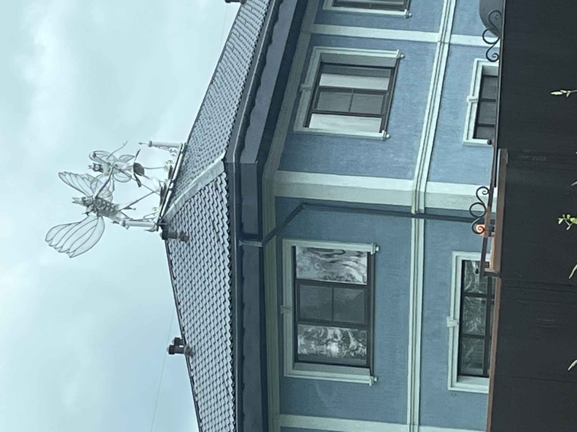 The image shows a two-story blue house with a dark gray tiled roof, located in Ol'gino, Russia. The roof is adorned with a metal sculpture of two winged figures, resembling fairies or elves, standing on a decorative architectural element. The house has several windows, some with white trim and darker frames, and the walls are painted a light blue with white vertical and horizontal accents. In the foreground, a dark brown fence is visible, with decorative metal scrolls on top. The sky is overcast and cloudy.