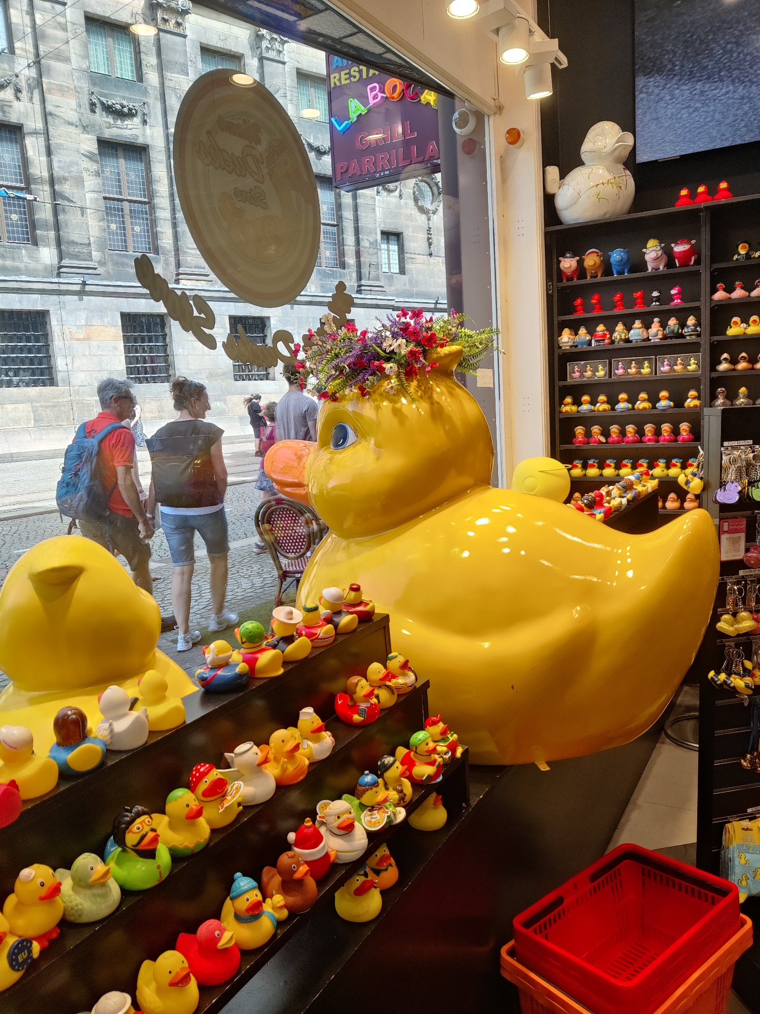 This is a photograph taken at the Duck Store in Amsterdam, Netherlands. The store is filled with a large collection of rubber ducks, including a giant yellow duck sculpture prominently displayed in the foreground, adorned with a floral arrangement on its head. Various shelves behind the sculpture are stocked with numerous smaller rubber ducks of different designs and colors, some arranged on display shelves and others in baskets. In the background, a window looks out onto a street scene with people walking by and a building facade with ornate architectural details. To the right of the shelves, a rack displays keychains with duck motifs. A red plastic basket sits in the lower right corner of the image, stacked on top of an orange bin.