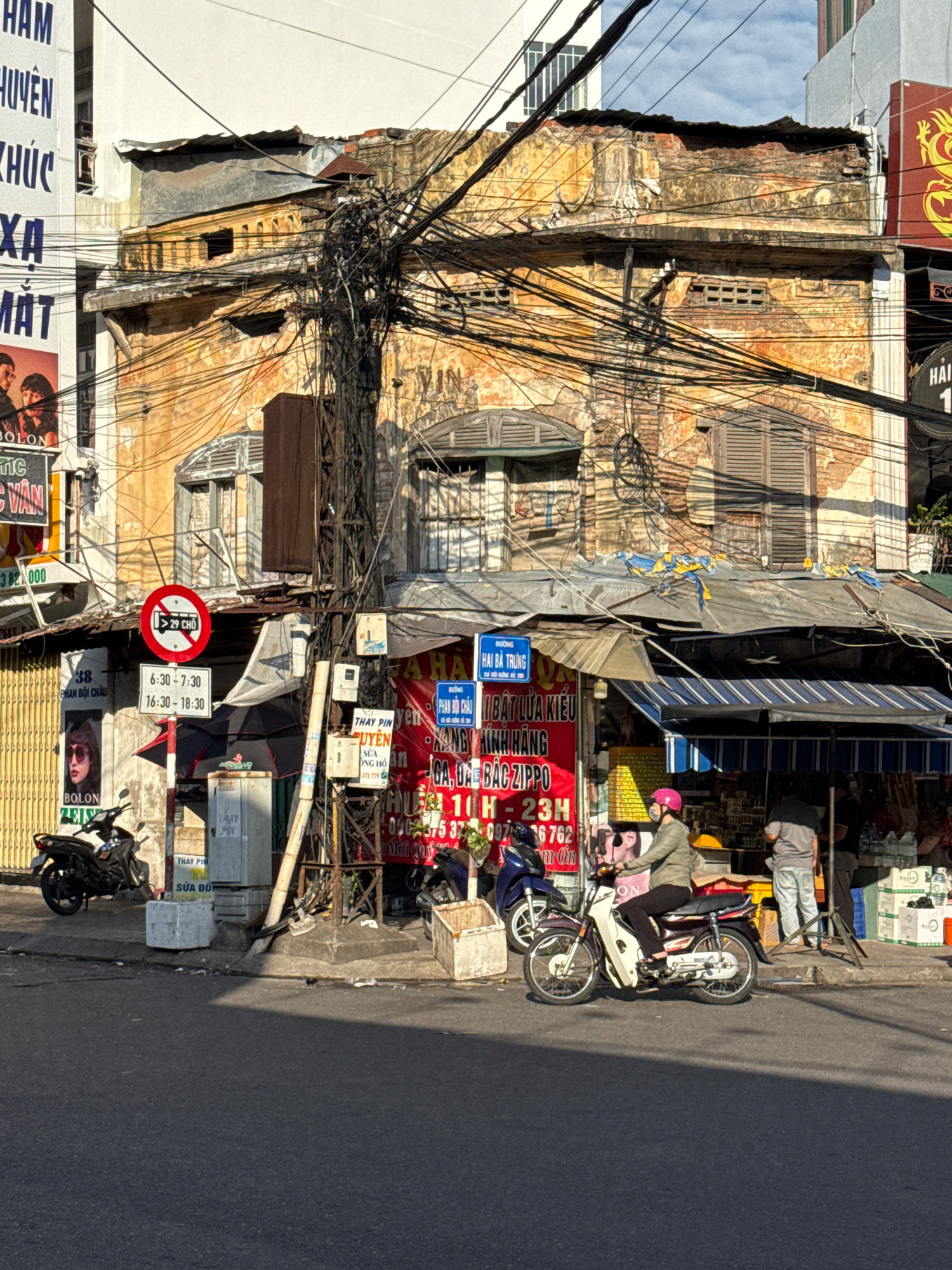 This is a street-level, eye-level photograph taken in Nha Trang, Vietnam. The scene is dominated by a weathered, two-story building with peeling yellow paint and extensive tangles of electrical wires. The building appears to be a shopfront with multiple signs, some in Vietnamese. One prominent red and white sign on the left has an image of a woman wearing sunglasses. A circular red and white traffic sign indicates a prohibition related to vehicles and a number "29 CHÔ". Below this, a schedule shows times in the format "6:30 - 7:30" and "16:30 - 18:30".
To the right of the traffic sign, a blue sign with Vietnamese text is mounted. In front of the building, a metal awning with blue and white stripes covers a stall or market area where several people are visible. A woman wearing a pink helmet and a green jacket is riding a white and maroon motorcycle towards the right side of the frame. Another person, wearing a grey shirt and light blue pants, stands near the market stall. A motorcycle is parked on the left side of the image. The street in the foreground is paved, with shadows cast by the buildings and overhead wires. The sky is visible in the upper portion of the image, appearing partly cloudy. The overall impression is one of a busy, somewhat chaotic urban environment.