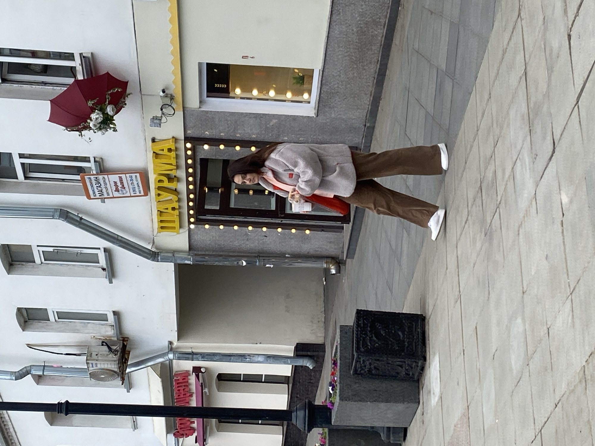 A young woman walks on a sidewalk in Moscow, Russia. She wears a light gray jacket, brown pants, and white shoes, carrying a red bag. The sidewalk is paved with gray rectangular stones. Behind her, a building facade is visible, painted white with multiple windows and a red umbrella decoration. A yellow sign with the word "SHAURMA" (a type of Middle Eastern street food) is prominently displayed. A black decorative structure stands on the sidewalk near the woman.