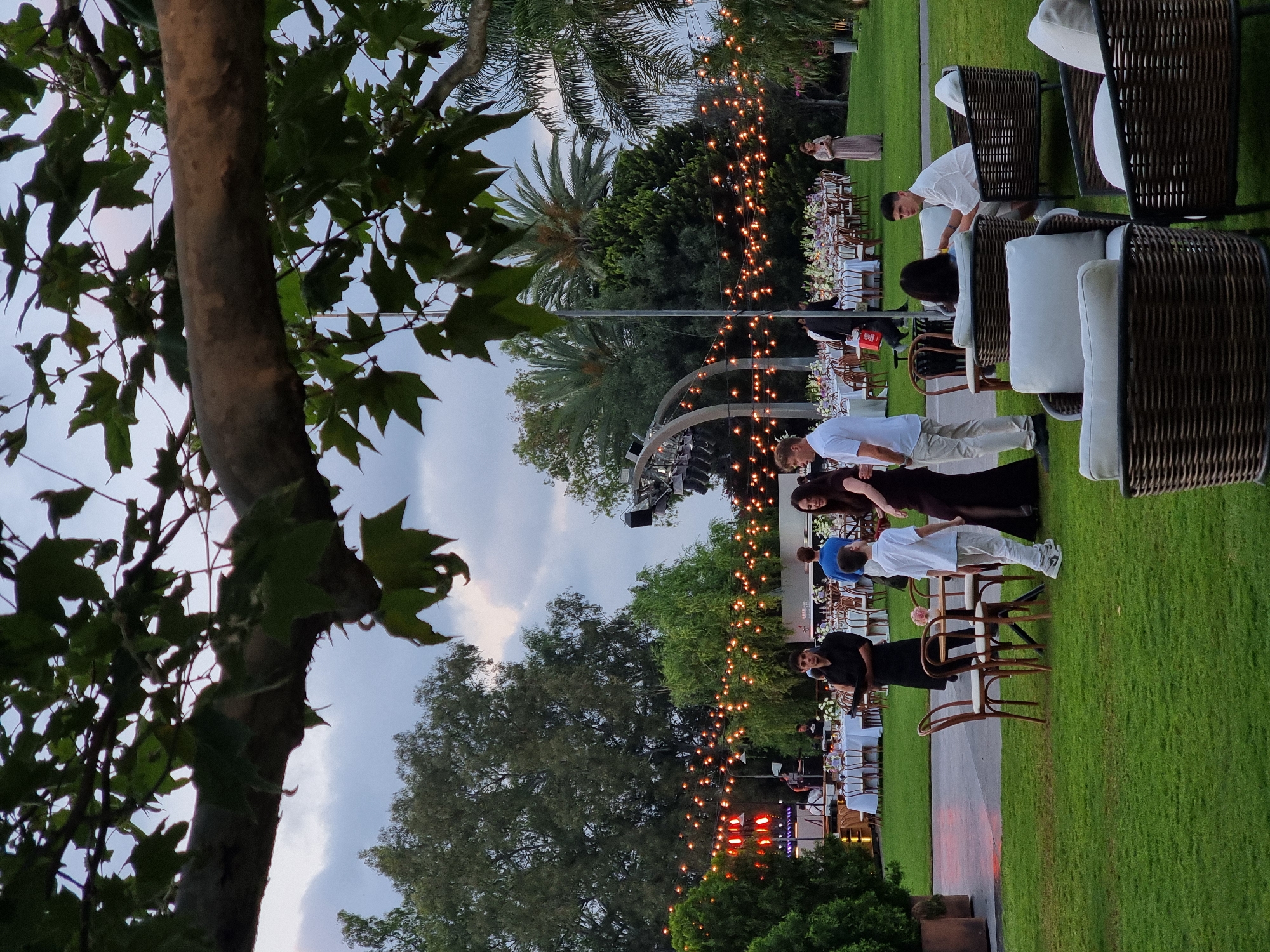 The image depicts an outdoor event, likely a wedding or formal gathering, held at the Ga'ash Golf Club in Ga'ash, Israel. The scene is set in the late afternoon or early evening, with ambient lighting provided by strings of incandescent bulbs stretched across the lawn and over a stage area.
In the foreground, a meticulously manicured green lawn is visible, with several wicker chairs and sofas arranged for seating. One sofa in the lower right corner has a person seated in it, facing away from the camera. Another sofa in the center right has a young man in a white shirt seated and facing left.
Further into the scene, a long, white-clothed table is set with chairs, indicative of a dining area. Several people are mingling around this area, including a woman in a dark dress and a young boy in a white shirt and pants, who appear to be interacting. A man in a black uniform, possibly a waiter or staff member, is holding a clipboard and walking past.
In the background, a stage with lights and sound equipment is set up, with a band or performers visible on it. Beyond the stage, trees and palm trees frame the outdoor venue. The sky is overcast with a mix of light and dark clouds. The overall atmosphere suggests a sophisticated outdoor event being held at dusk.