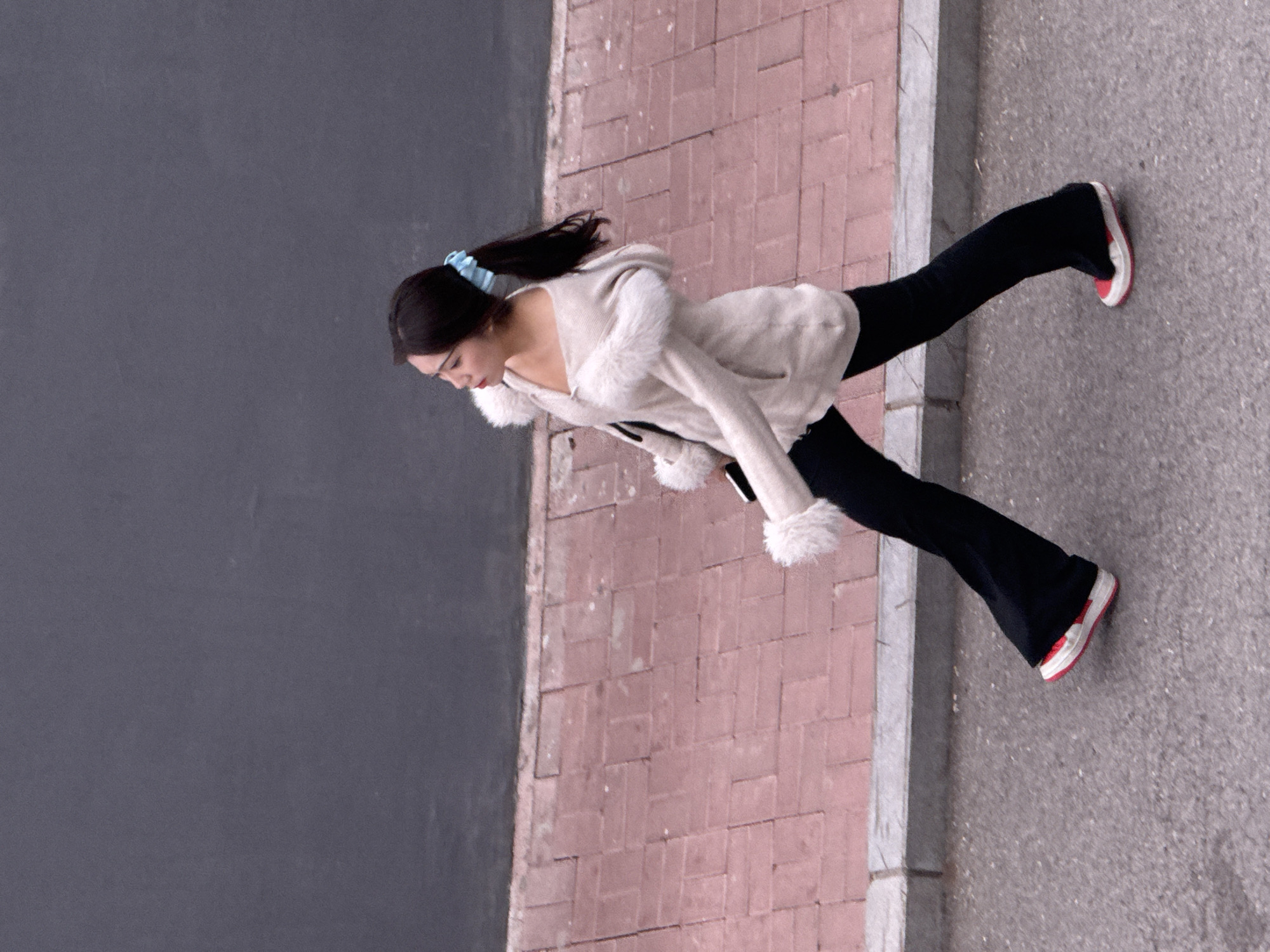 A high-angle shot depicts a young woman walking along a sidewalk in what is identified as the city of Zhongtun, China. The woman has dark hair pulled back with a blue scrunchie. She is wearing a cream-colored, fur-trimmed hooded sweater and black pants, and her footwear consists of red and white sneakers. She is holding a mobile phone. The setting includes a dark gray wall to her left, and a brick-paved area next to the sidewalk.