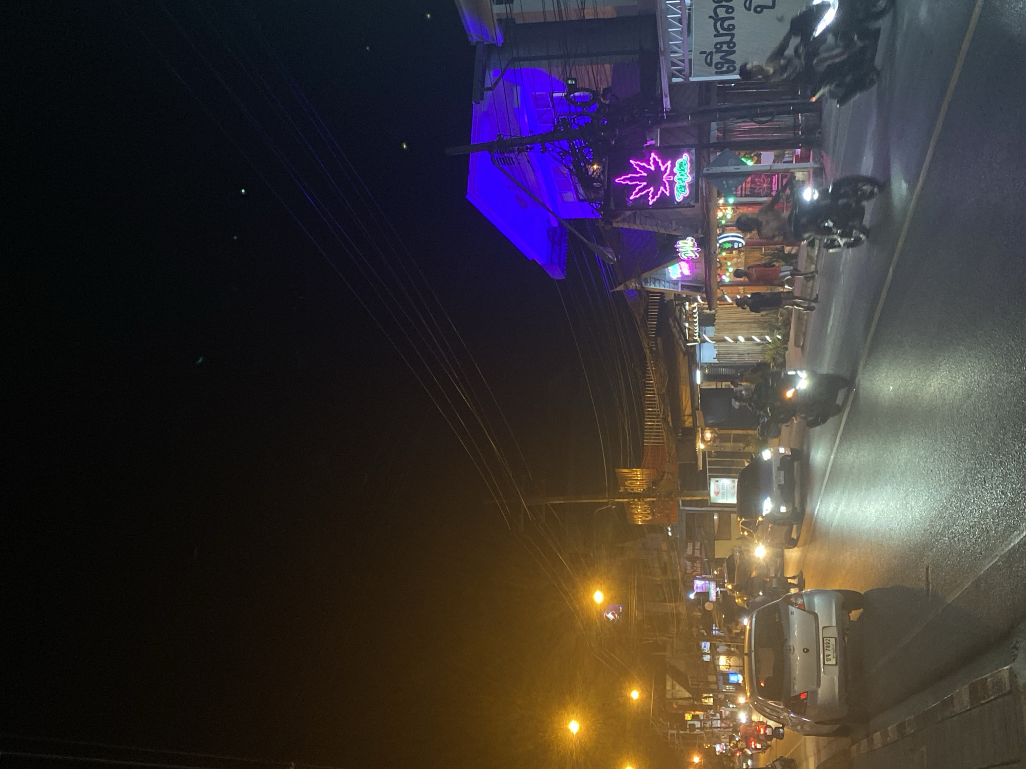 The image captures a street scene at night in Baan Mae Nam, Thailand. A street runs from the bottom right to the top center, wet from recent rain, with traffic including motorcycles and cars. A silver car is parked on the right side of the street. Shops line the left side of the street, illuminated with colorful neon lights, including a sign with a cannabis leaf. People are walking along the sidewalk in front of the shops. Overhead wires cross the dark sky above the scene. Streetlights provide additional illumination along the street.