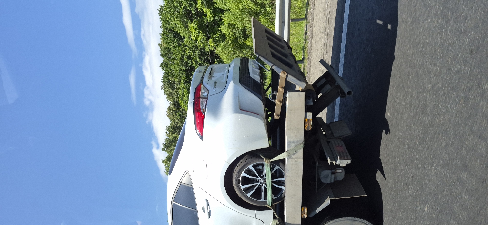 A white sedan is being transported on the back of a tow truck on a sunny day. The car is secured to the flatbed with green straps. The tow truck is driving on a paved road, with a guardrail and trees lining the right side of the road. The sky is blue with scattered white clouds. The scene appears to be on a highway outside of the city of Gorodechnoe, Russia.