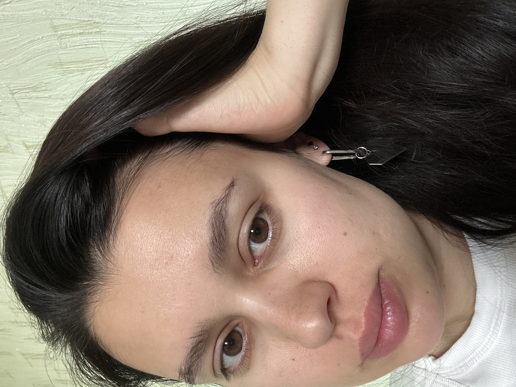 A close-up, slightly rotated view of a woman's face. She has dark brown, straight hair that appears to be pulled back or to the side, revealing a portion of her forehead. Her skin is fair, and she has brown eyes. A silver earring with a rectangular pendant is visible in her ear. Her lips are full and pink. Part of a hand is visible above her hair, suggesting she is touching it. The background is a textured, light-colored surface. There is no indication of the location or city within the image.