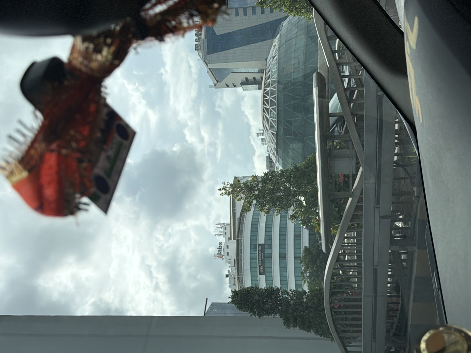 The image captures a view from within a vehicle looking out at a cityscape in Hyderabad, India. In the foreground, a part of the vehicle's interior, including a portion of the dashboard and windshield, is visible. A decorative item, possibly a car accessory, hangs from the rearview mirror, partially obscuring the sky.
Dominating the mid-ground are several modern buildings. On the left, a curved white building with multiple floors and numerous windows is prominent, displaying signage that reads "JUBS EDUCATION INDIA". Above this building, the "JUBS" logo is visible. To the right of this building, a large, geodesic dome structure with a metal framework is situated. Behind the dome, a tall, angular glass-fronted building stands, with signage that appears to read "BNKI".
In front of the buildings, a pedestrian bridge with a curved railing and vertical supports spans across the frame. Trees and some flowering plants are visible on and around the bridge.
The sky is overcast with a mix of clouds, indicating a potentially cloudy or rainy day. The overall impression is of a contemporary urban environment with a blend of architectural styles. The specific location is likely a commercial or business district within Hyderabad, India.