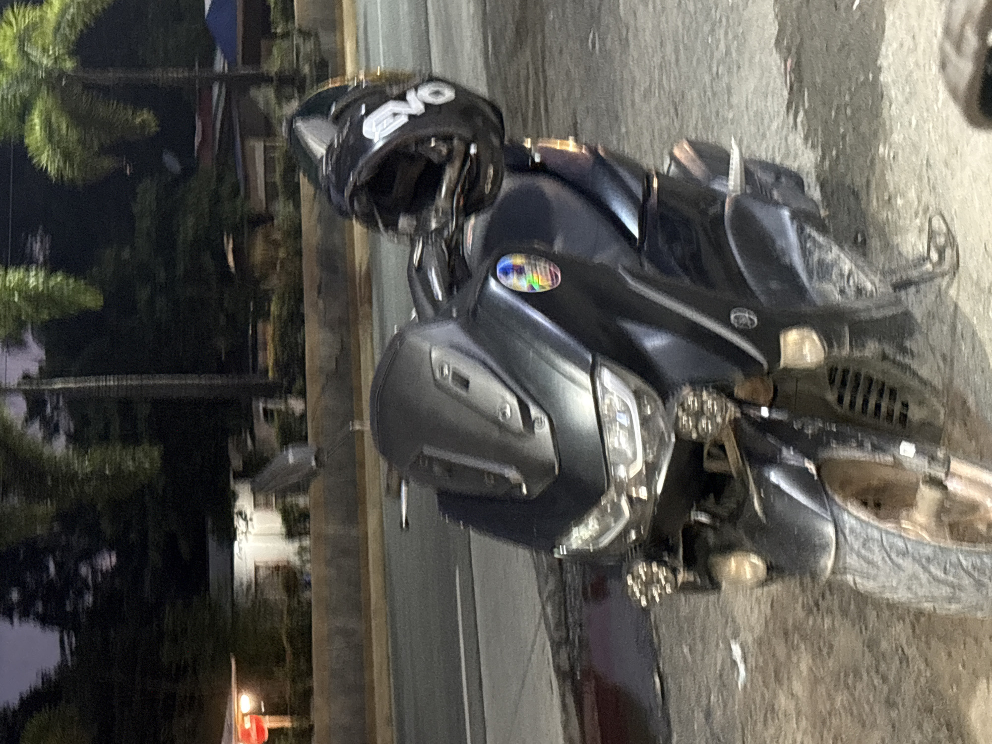 A black scooter is parked on a dirt road next to a paved road. A black helmet with the word "EVO" is resting on the handlebars of the scooter. In the background, palm trees and buildings are visible. The scene appears to be a roadside location in Carmen, Philippines, based on the distinctive style of the buildings and vegetation. The lighting suggests it is evening or nighttime.