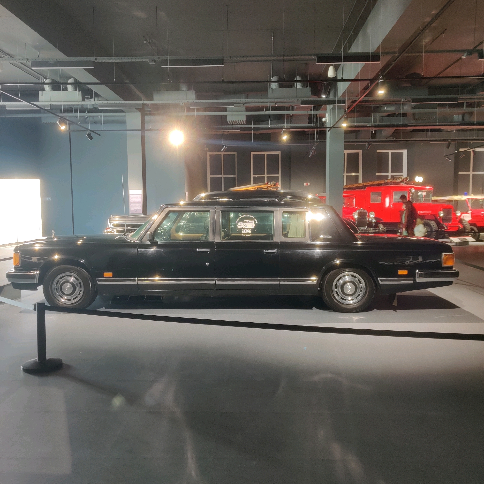 The image captures an indoor exhibition space in the city of Verkhnyaya Pyshma, Russia. In the foreground, a long, black limousine is prominently displayed. The vehicle exhibits a classic, elongated design with multiple windows along its side. A shiny chrome trim runs along the lower portion of the car's body. The limousine rests on a grey surface, with a black stanchion and rope barrier visible in the lower left corner, indicating a protected exhibit.
In the background, several other vehicles are partially visible. To the right, a bright red fire truck is parked, featuring large wheels and an open cabin. Behind the fire truck, another vehicle, possibly another truck or car, is also present. The environment appears to be a museum or exhibition hall, characterized by a dark ceiling with exposed industrial elements, such as conduits and lighting fixtures. Spotlights illuminate various areas of the display. A person, partially obscured, is visible near the red fire truck. The overall lighting is a mix of dramatic spotlighting and ambient illumination, creating reflections on the polished surfaces of the vehicles. The wall behind the limousine is a muted blue-grey.