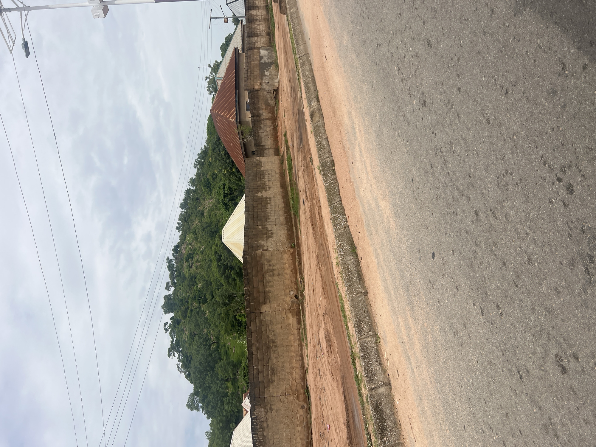 This is a landscape photograph taken from the side of a paved road in Minda, Nigeria. The road appears to be in fair condition with some visible patches and stains. A concrete curb separates the road from a dirt path, which runs alongside a brick wall. Beyond the wall, the terrain slopes upwards, covered in lush green vegetation and trees. The sky is overcast with scattered clouds, suggesting it might be a cloudy day.
In the background, several buildings with corrugated iron roofs are visible, nestled against the hillside. There are also power lines stretching across the frame, indicating the presence of infrastructure. The overall scene depicts a rural or semi-urban environment in Nigeria, characterized by a mix of natural landscape and man-made structures. There is no visible text in the image, nor any clear indication of a specific activity or event taking place, other than it being a typical day in this location. The lighting suggests it is daytime, but the overcast sky limits strong shadows, creating a diffused light. The image evokes a sense of everyday life in a Nigerian setting, with the natural environment playing a prominent role.