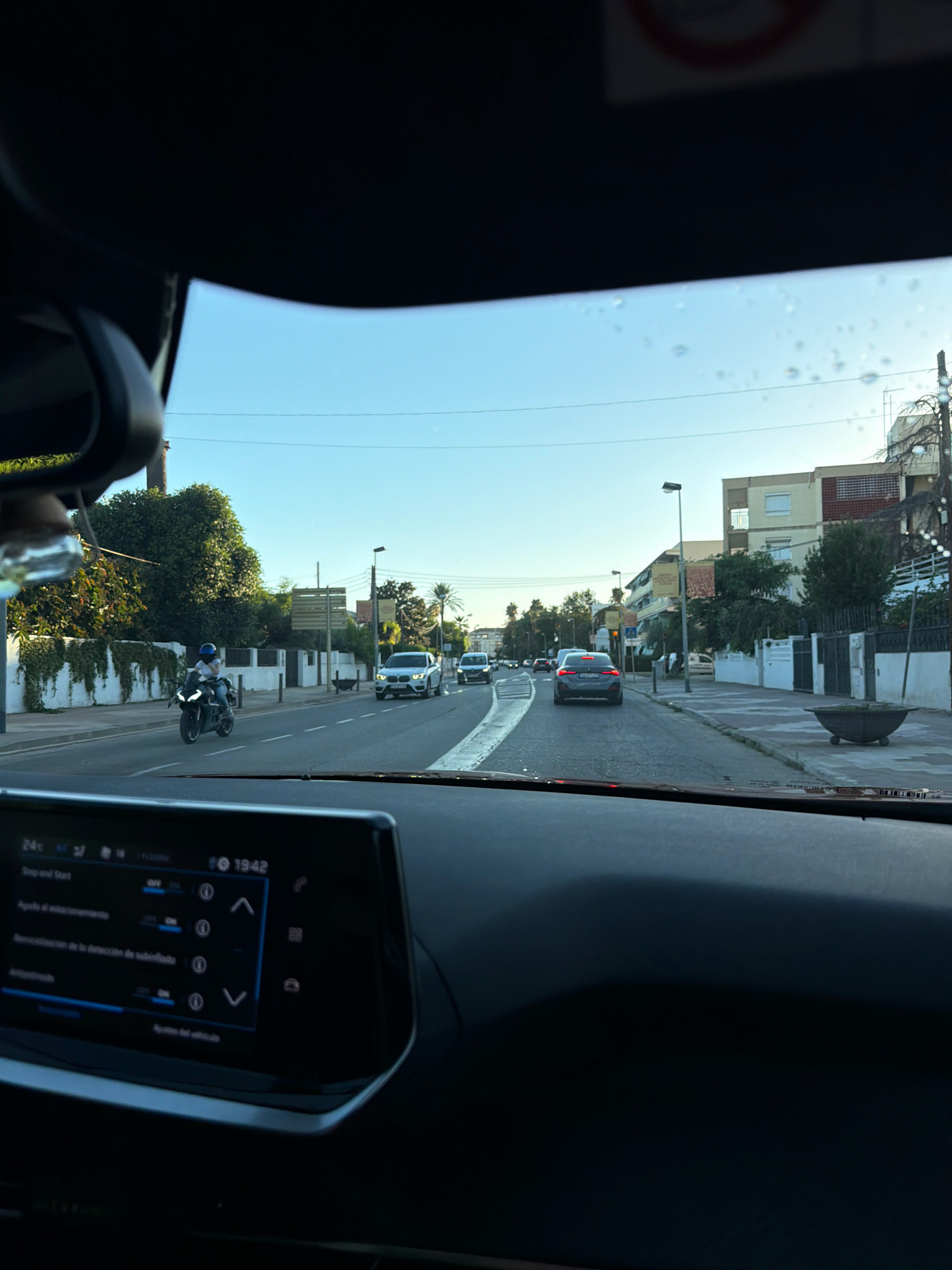 A view from inside a car in the city of Vilafortuny, Spain. The perspective is looking forward through the windshield. The sky is clear and blue. A road with traffic stretches ahead, lined by buildings and trees. Several cars and a motorcycle are visible on the road. The dashboard of the car is in the foreground, displaying a digital interface. The scene appears to be a street in a residential or mixed-use area, possibly during daytime.
