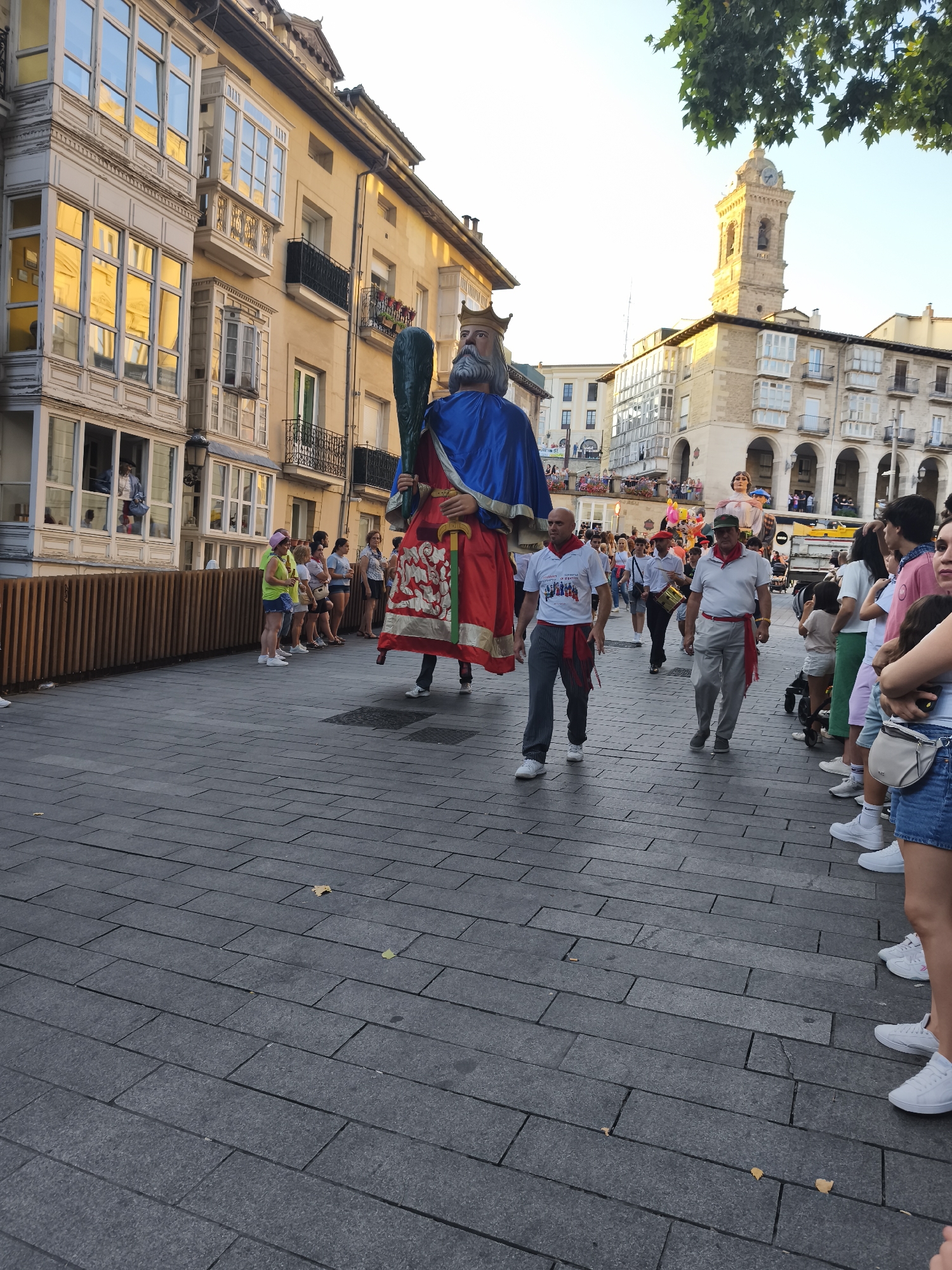 The image depicts a street scene in Vitoria-Gasteiz, Spain, likely during a cultural event or parade. A large, costumed figure, possibly a "Gigante" (giant) character from local folklore, dominates the foreground. The giant is adorned in a blue cape and a red and gold tunic, wearing a crown and holding a scepter. It appears to be moving down a pedestrianized street paved with dark grey rectangular stones.
A group of people are walking alongside and behind the giant. Some are dressed in casual attire, while others wear t-shirts with a red sash or band around their necks, suggesting participation in the event. One man in the foreground wears a white t-shirt with a graphic design and dark striped pants. Another man, also in the foreground, is dressed in a white polo shirt, grey pants, and a red sash. Behind them, a person in a red beret appears to be drumming.
To the left of the street, a multi-story building with ornate balconies and large windows lines the street. The windows reflect the golden light of what appears to be late afternoon or early evening. To the right, more buildings with arched structures and balconies are visible, and a clock tower is prominent in the background.
A crowd of spectators is gathered on the right side of the street, observing the procession. They are of various ages, and some are dressed in summer clothing, including shorts and t-shirts. The overall atmosphere suggests a festive and public gathering. A low wooden fence or barrier separates the street from the sidewalk where some of the spectators are standing.