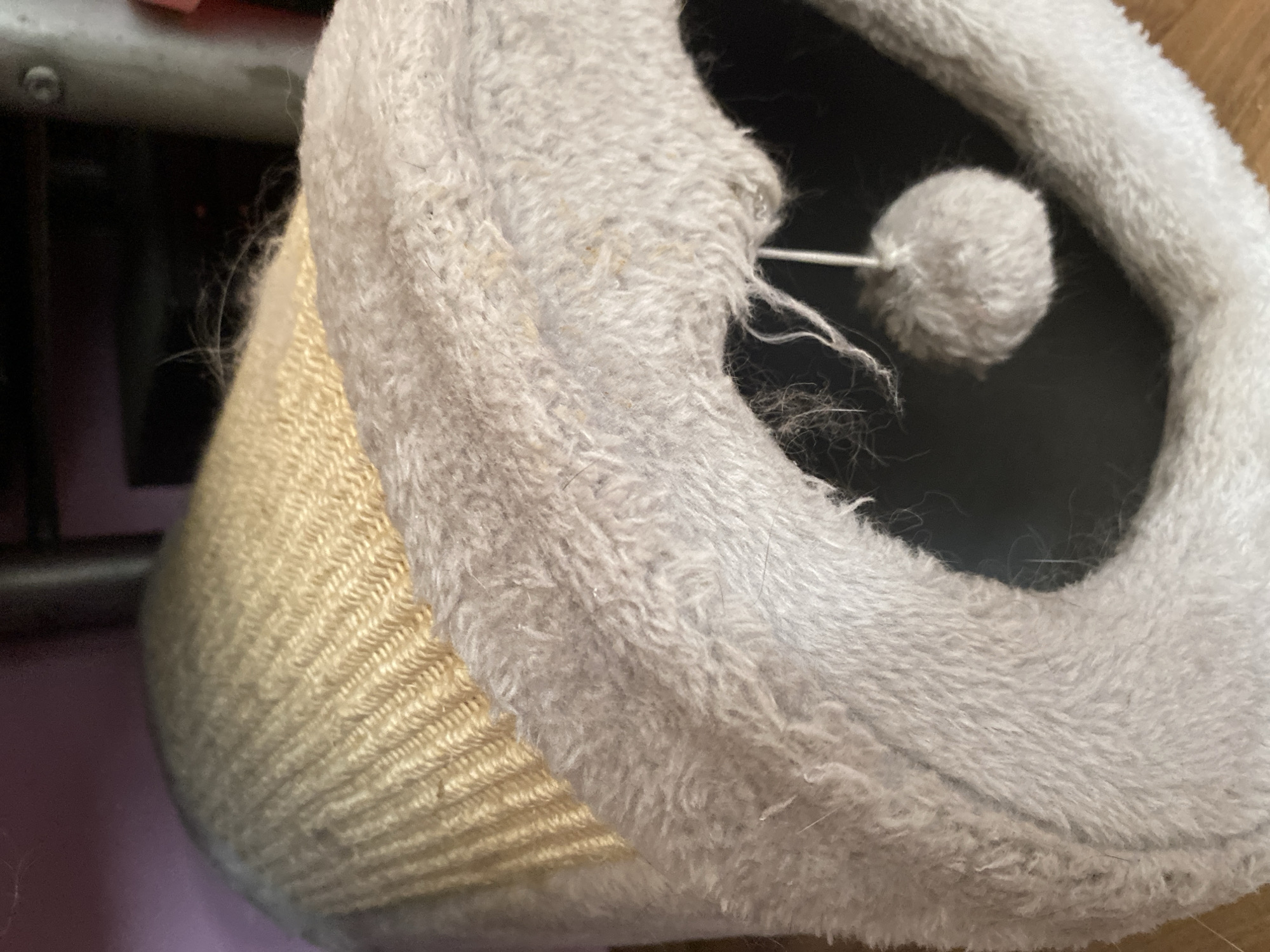 The image is a close-up, eye-level shot of a beige, woven scratching post with a grey plush-covered opening. The scratching post is cylindrical and appears to be made of sisal rope. A small, white pom-pom toy hangs by a string inside the plush opening. The background is out of focus and shows a dark, indistinct object and a faint pinkish-purple surface. There are no people present in the image. The scene is indoors, and there is no information to suggest a specific location within the city of Bílina, Czechia.