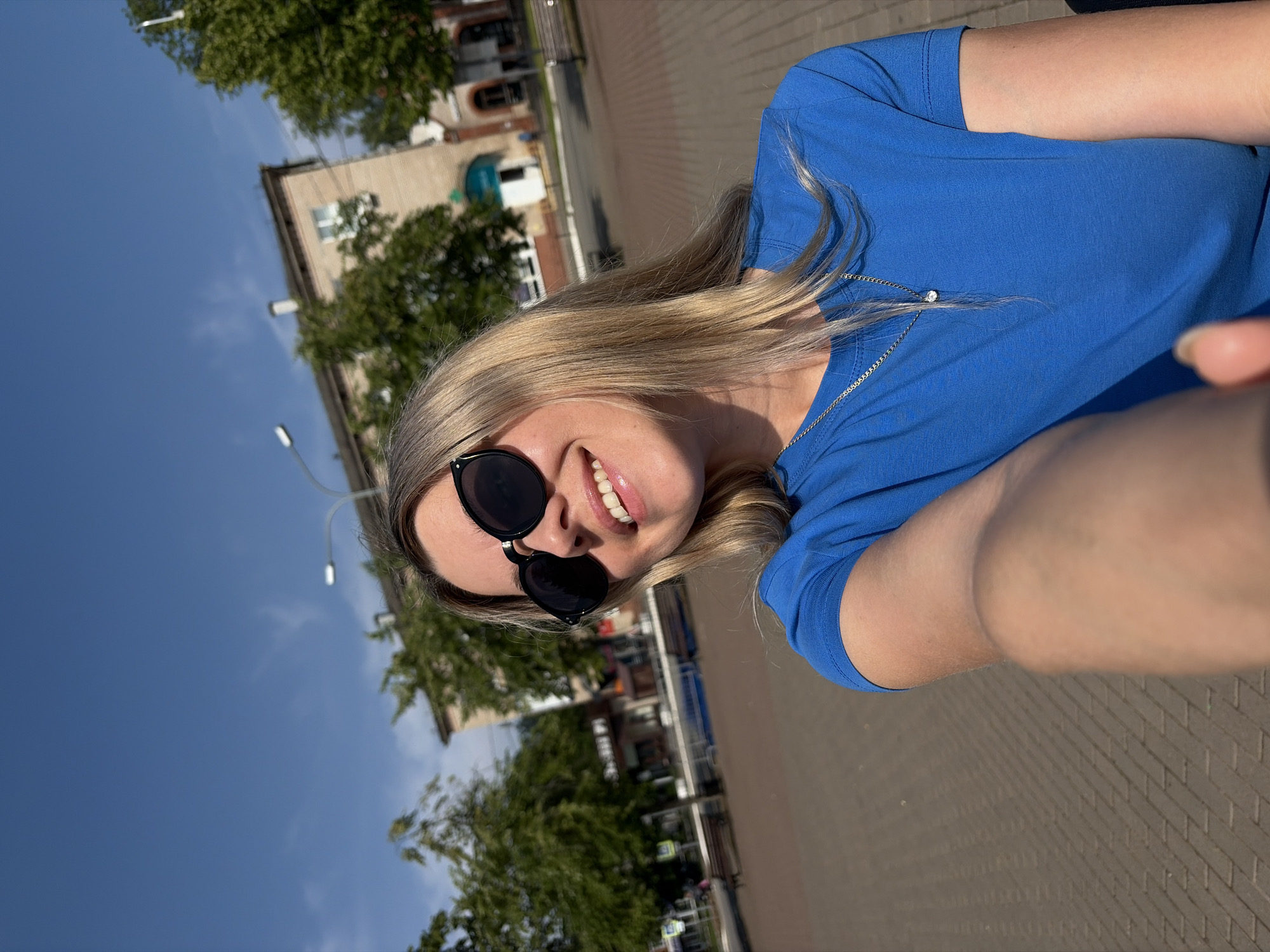 A blonde woman wearing a blue t-shirt and sunglasses smiles while taking a selfie outdoors. She is in an urban environment with buildings and trees visible in the background. The ground is paved with bricks. The sky is clear and blue. The location appears to be the city of Chaikovsky, Russia, possibly near a central square or park.