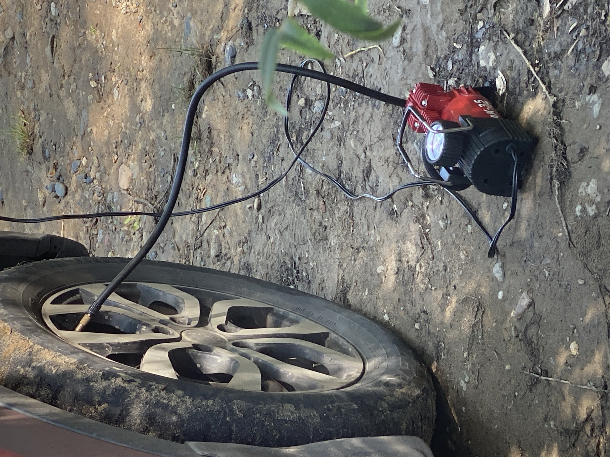 A close-up shot captures a portable tire inflator connected to the valve stem of a car tire. The inflator, a compact red and black device, sits on the dusty, uneven ground beside the vehicle. A black hose extends from the inflator to the tire's valve. The car tire, partially visible, appears to be dirty. The background consists of dry earth, small stones, and sparse vegetation, suggesting an outdoor, possibly rural or unpaved area. The lighting indicates it is daytime. No people are present in the frame. The scene appears to be in Novokuznetsk, Russia.