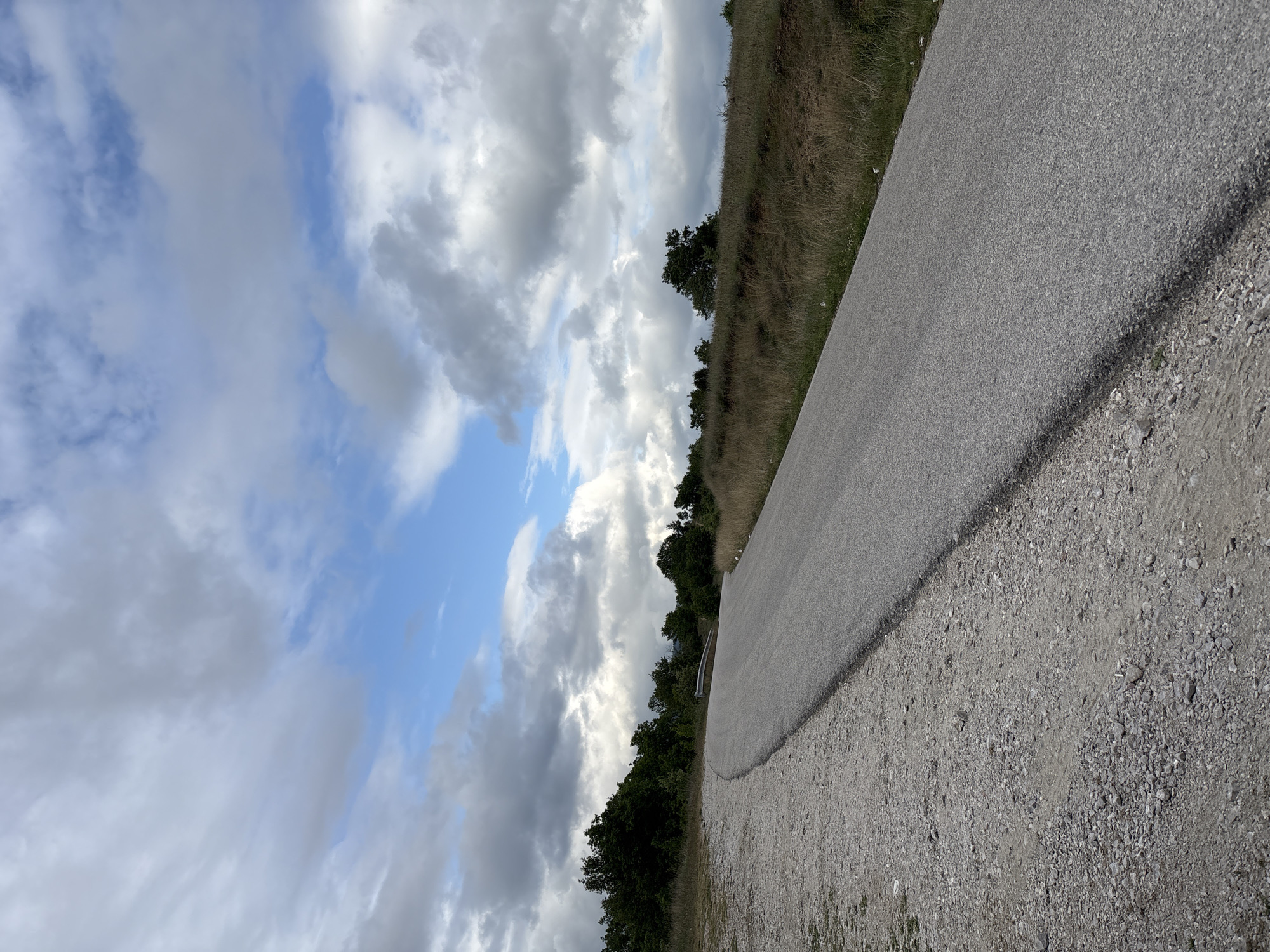 A ground-level shot captures a paved road, gray in color, occupying much of the right side of the frame. The road is flanked by loose gravel and vegetation. To the left of the road, there is a line of trees and shrubs. Above them is a sky filled with a mix of white and gray clouds, with patches of blue visible through the breaks in the cloud cover. The image appears to be taken in daylight. No people or interactions are visible in the scene. The location is unknown.
