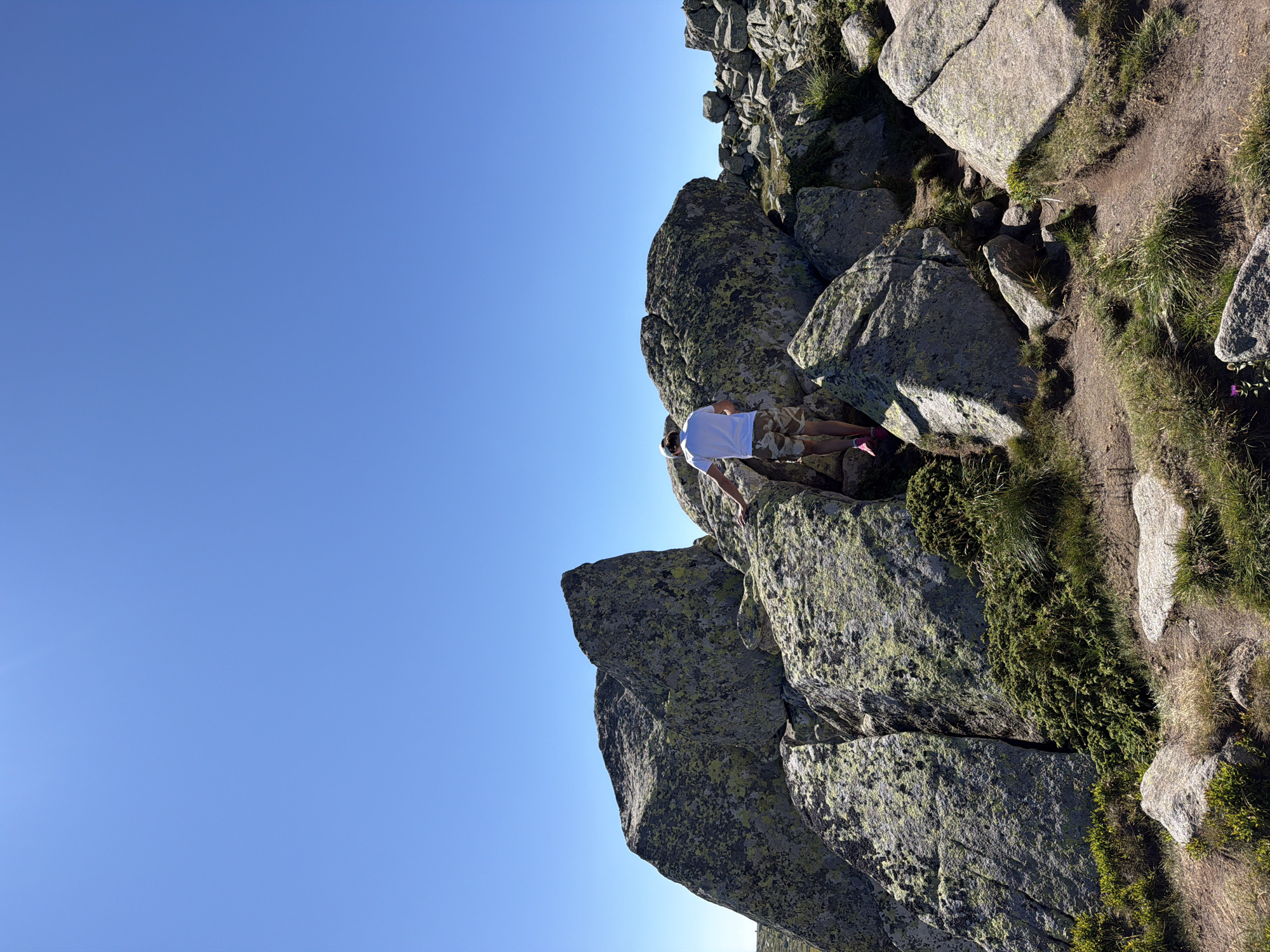 The image shows a person walking on a rocky terrain under a clear blue sky. The person is wearing a white t-shirt, camouflage shorts, and pink shoes. They are positioned in the center-right of the frame, facing away from the camera and ascending over large, lichen-covered rocks. The landscape is characterized by rough, uneven ground with scattered vegetation. The scene appears to be a mountain or high-altitude area, potentially within the vicinity of Bistritsa, Bulgaria, known for its natural landscapes and hiking trails. The sunlight is bright, casting shadows on the rocks.