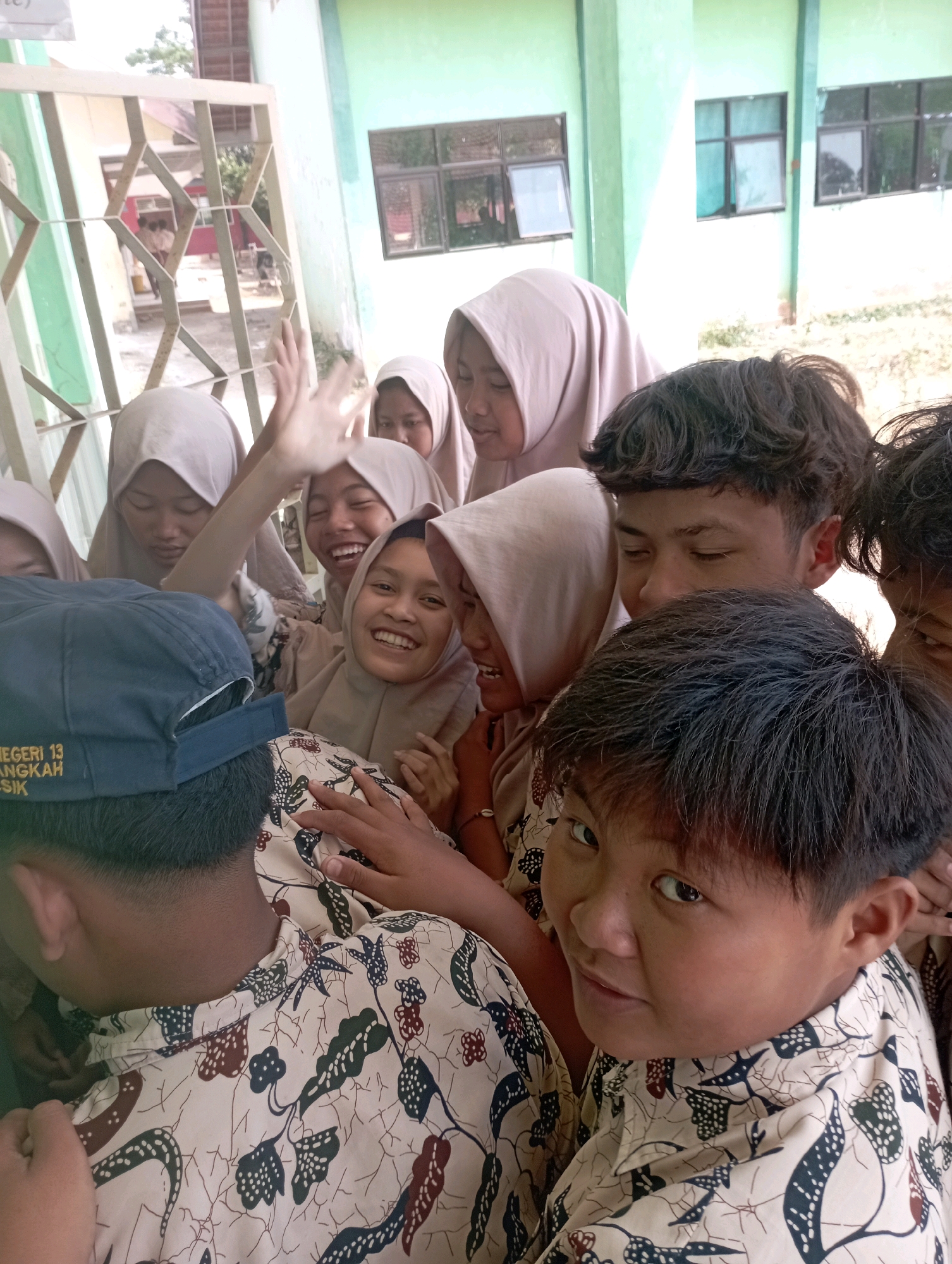 In Banyuurip, Indonesia, a group of school-aged individuals, some wearing hijabs, are gathered outdoors in front of a light green building with windows. The individuals are close together, with their faces visible and some smiling. One person in the foreground wears a dark blue baseball cap with yellow text that reads "NEGERI 13 LANGKA" and "SIK". Another individual in the foreground is looking directly at the camera with a slight smile. The people are wearing patterned shirts. The background shows a fence and more of the school building.