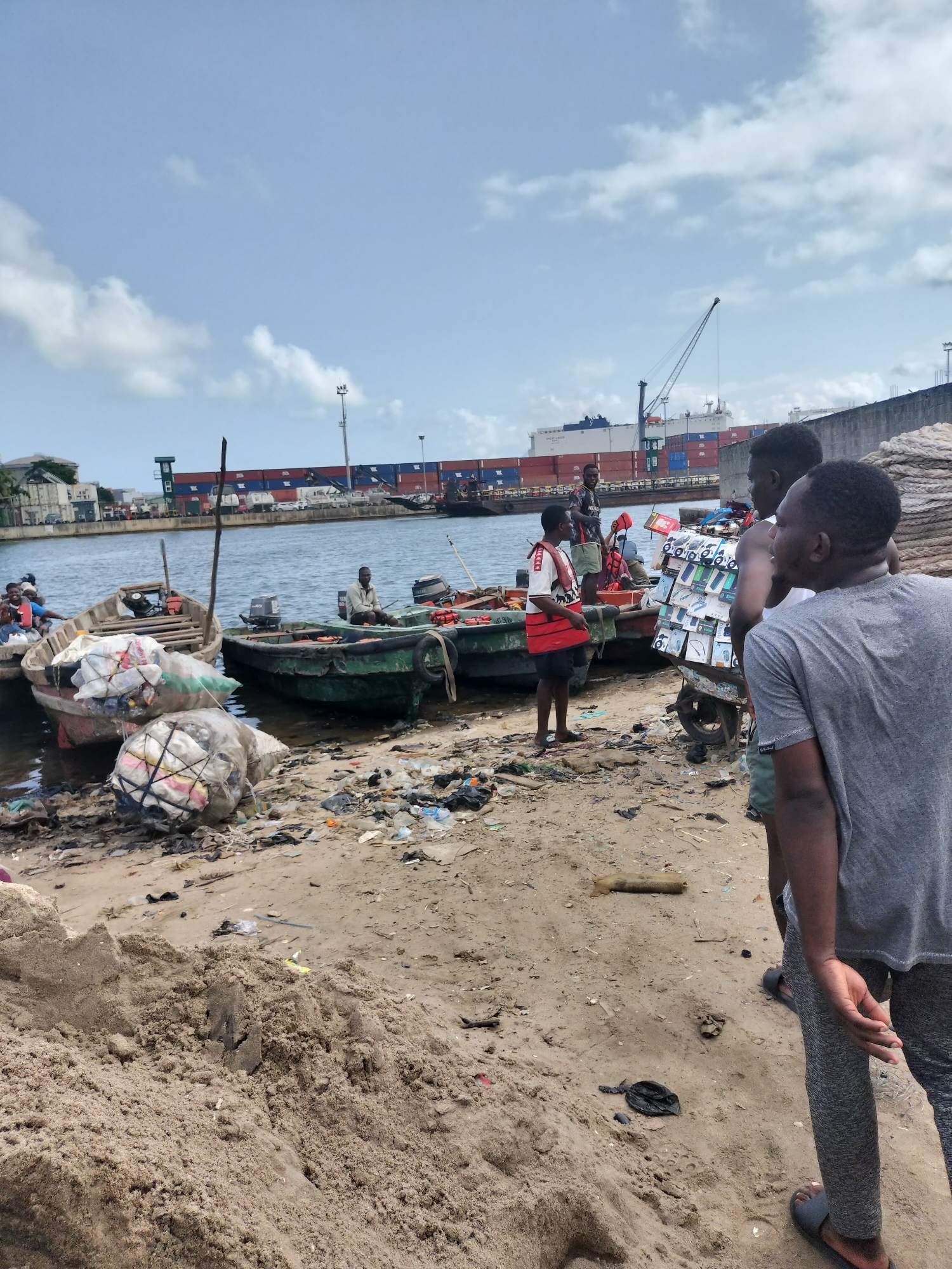 The image captures a bustling, somewhat cluttered waterfront scene in Alaiyabiagba, Nigeria, under a partly cloudy daytime sky. The foreground is dominated by sandy ground, with scattered piles of sand and a significant amount of litter, including plastic bags and debris.
Several people are visible in the midground and foreground. In the immediate foreground, a man with his back to the camera, wearing a grey t-shirt and grey pants with black sandals, looks towards the water. Next to him, another man with a bare torso pushes a two-wheeled handcart heavily laden with numerous small, packaged goods, possibly electronics or phone accessories. Near the water's edge, another man in a white and red striped shirt with an orange life vest stands beside a small wooden boat. Another man in a dark shirt is also by the boats. Further into the water, a man sits in a green boat equipped with an outboard motor.
The boats, predominantly small wooden vessels, are moored along the sandy bank. One boat contains large white mesh bags filled with what appears to be collected materials or cargo. The green boat also has similar bags. The water itself shows reflections of the sky and structures.
In the background, a wide body of water separates the immediate shore from a more industrial area. A very large container ship, painted white and laden with numerous red and blue shipping containers, dominates the distant horizon. Part of the ship's name "GOLD LAGO" and "LAGOS" are faintly visible on its side and containers. A large port crane and other industrial structures, including concrete walls, are also visible along the far bank, indicating an active port or shipping lane. The overall scene suggests a vibrant area of informal commerce and transport alongside heavy industry.